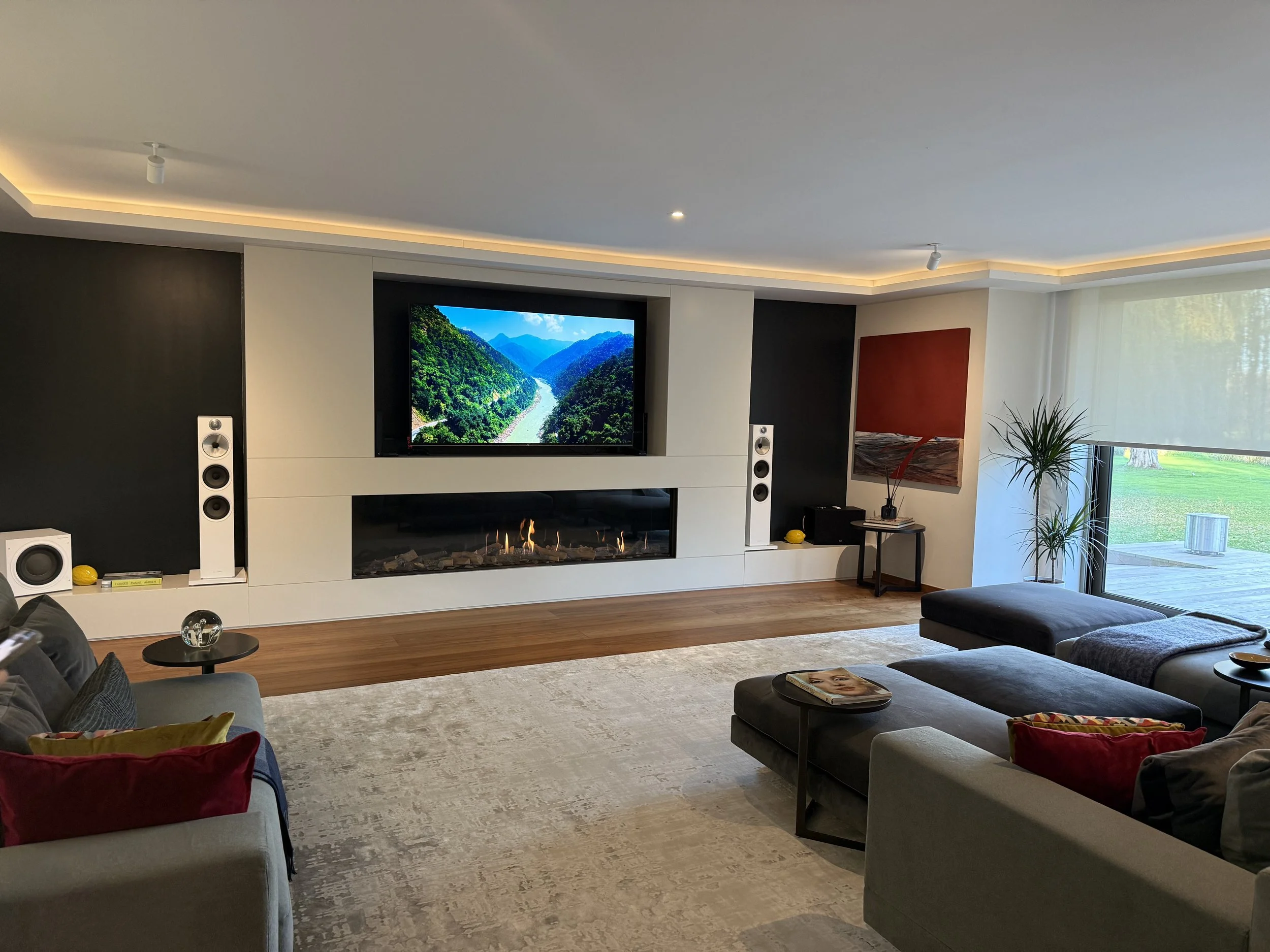 A modern living room featuring a wall-mounted TV displaying a mountain river scene, a fireplace beneath the TV, two tall white speakers on either side, a gray sectional sofa with colorful pillows, a small black side table, and a large window with whi