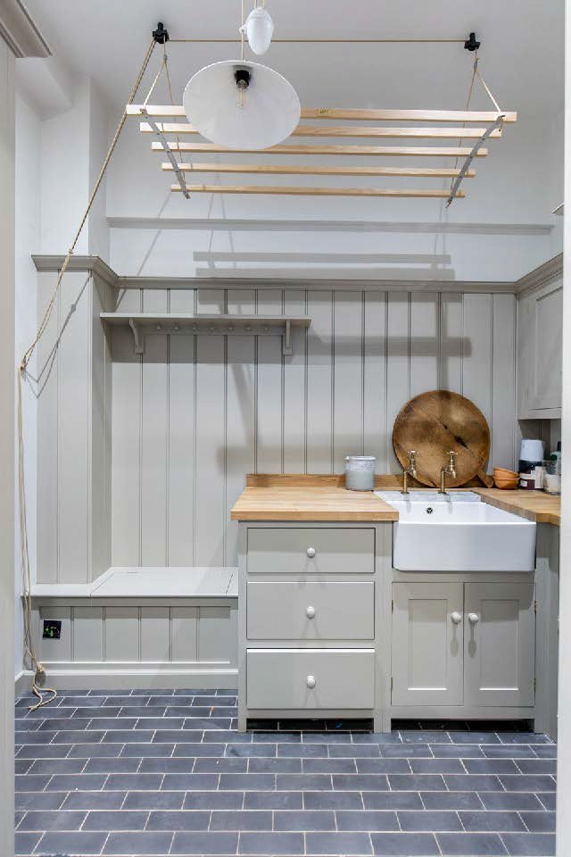 A small kitchen with light gray paneling walls, a white farmhouse sink, wooden countertops, and a gray cabinet with three drawers. There is a brass tray, a container, and other small items on the counter. Above, a ceiling-mounted wooden drying rack w