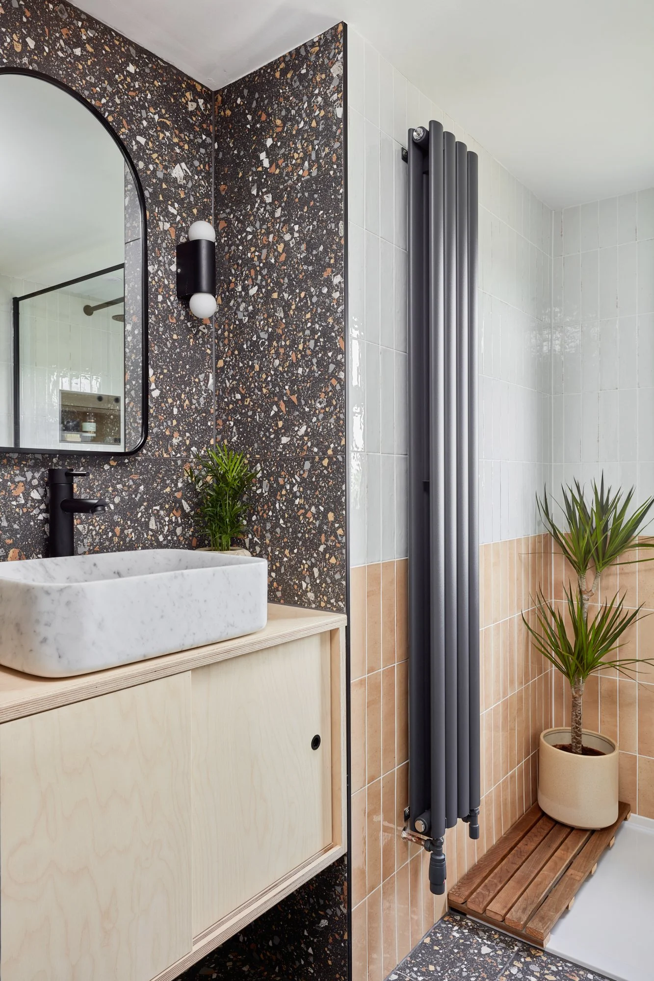 Modern bathroom with a terrazzo backsplash, a vessel sink, a black tap, a mirror, a black wall light, a tall dark gray radiator, a potted plant, and a wooden bench with a potted plant.