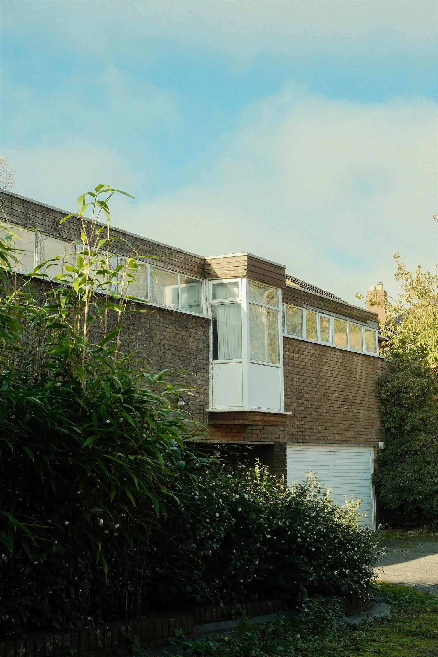 A two-story brick house with a white garage door, large front windows, a small balcony with white railing, and overgrown bushes and trees in front.