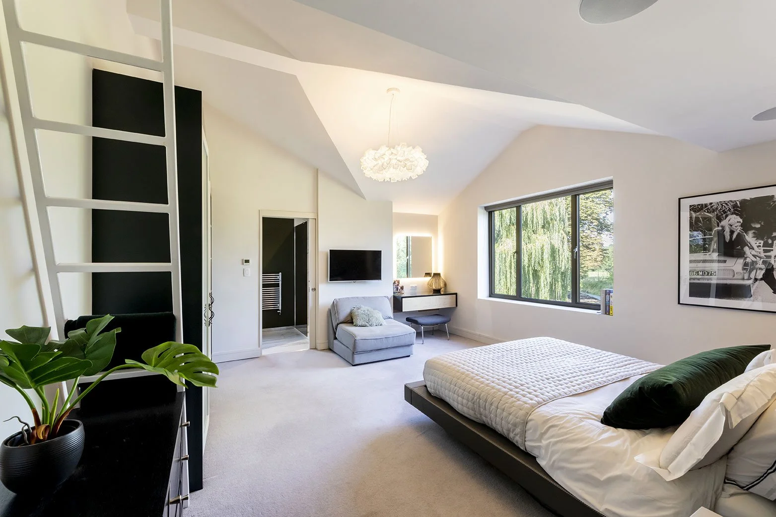 Modern bedroom with a large bed, black and white wall art, a window showing greenery outside, a gray chair, a small vanity with a mirror, a TV, and a plant in a black pot.