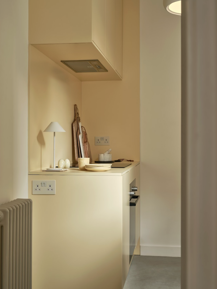 A small kitchen corner with cream-colored cabinets, a built-in oven, and a countertop holding decorative items like bowls, a lamp, and cutting boards. There are electrical outlets on the wall.