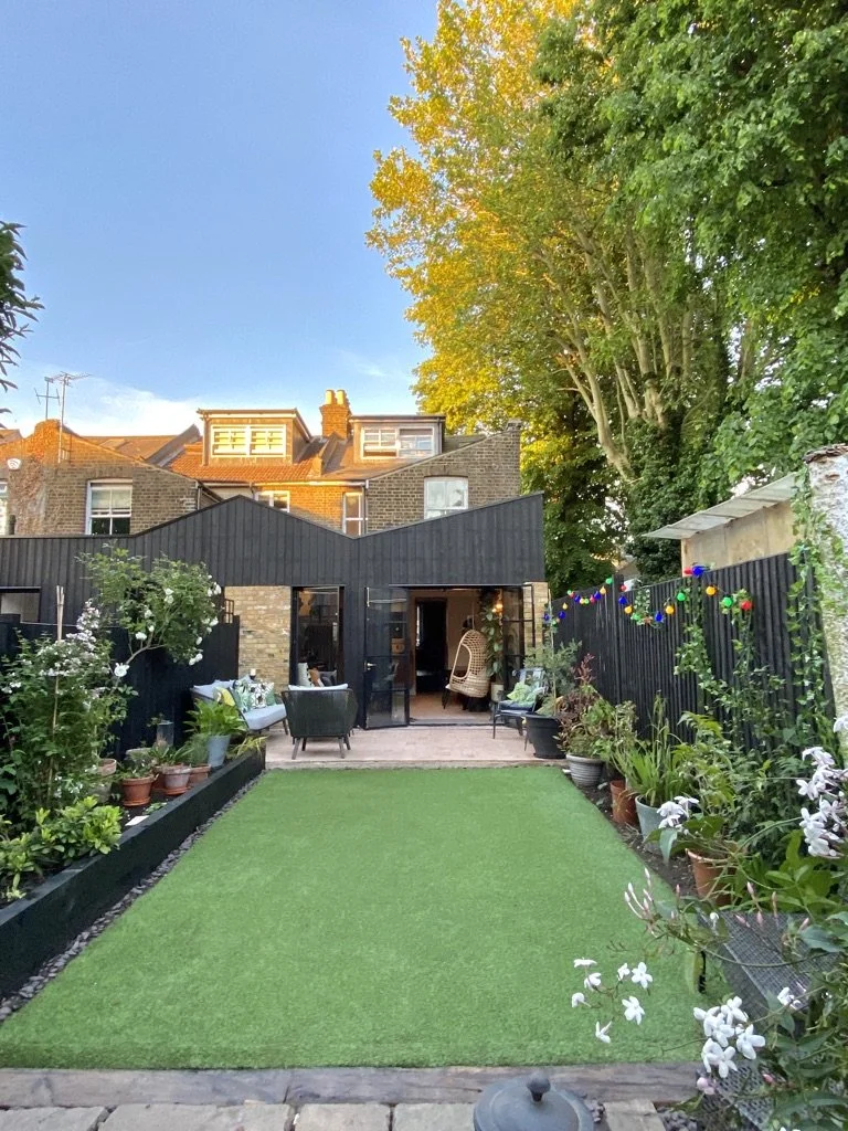 A backyard with a well-maintained green lawn, surrounded by potted plants and flowering bushes, with an outdoor seating area and string lights, and a black shed or building at the back.