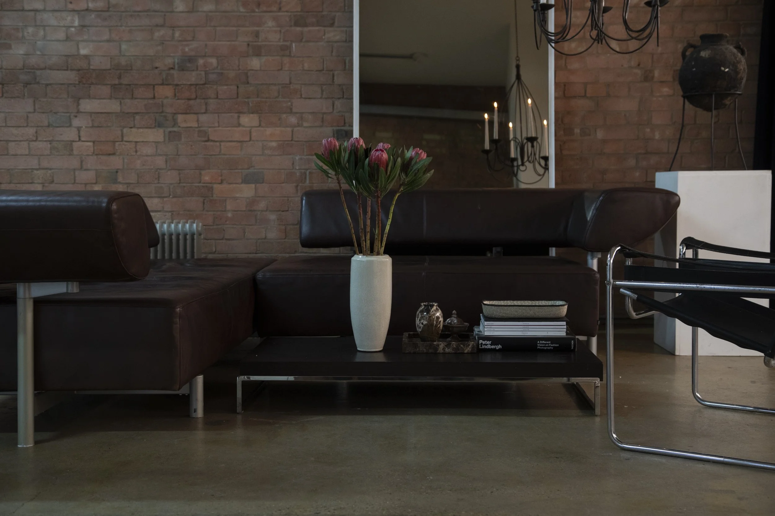 Interior of a living space featuring a brick wall, a black leather sectional sofa, a stack of books, a white vase with pink flowers, and decorative items on a black coffee table. Chandelier and mirror are reflected in the background.