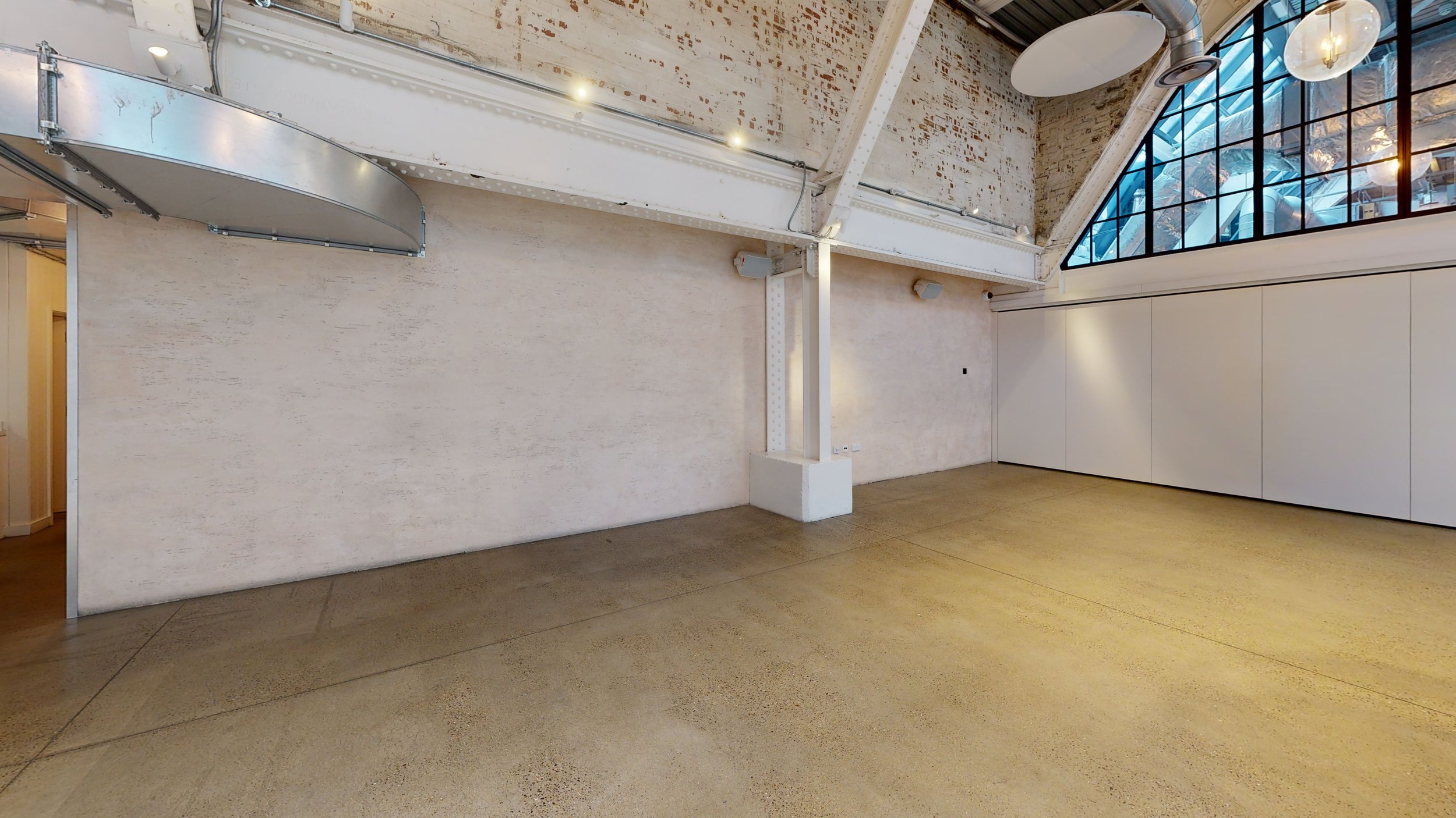 Empty interior space with beige polished concrete floor, exposed brick and beige painted walls, and large industrial-style window with a curved roof above.