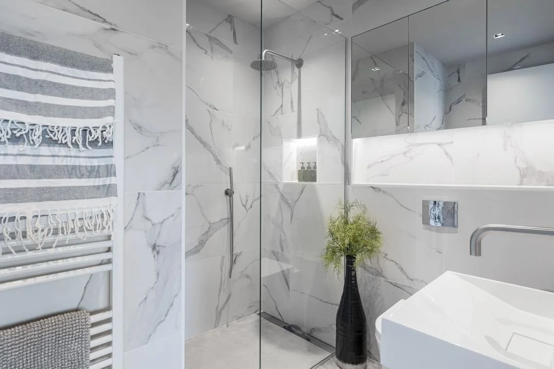 Modern bathroom with marble tiles, a walk-in shower with a glass door, a white towel on a wall-mounted radiator, a black vase with greenery, and a large mirror.