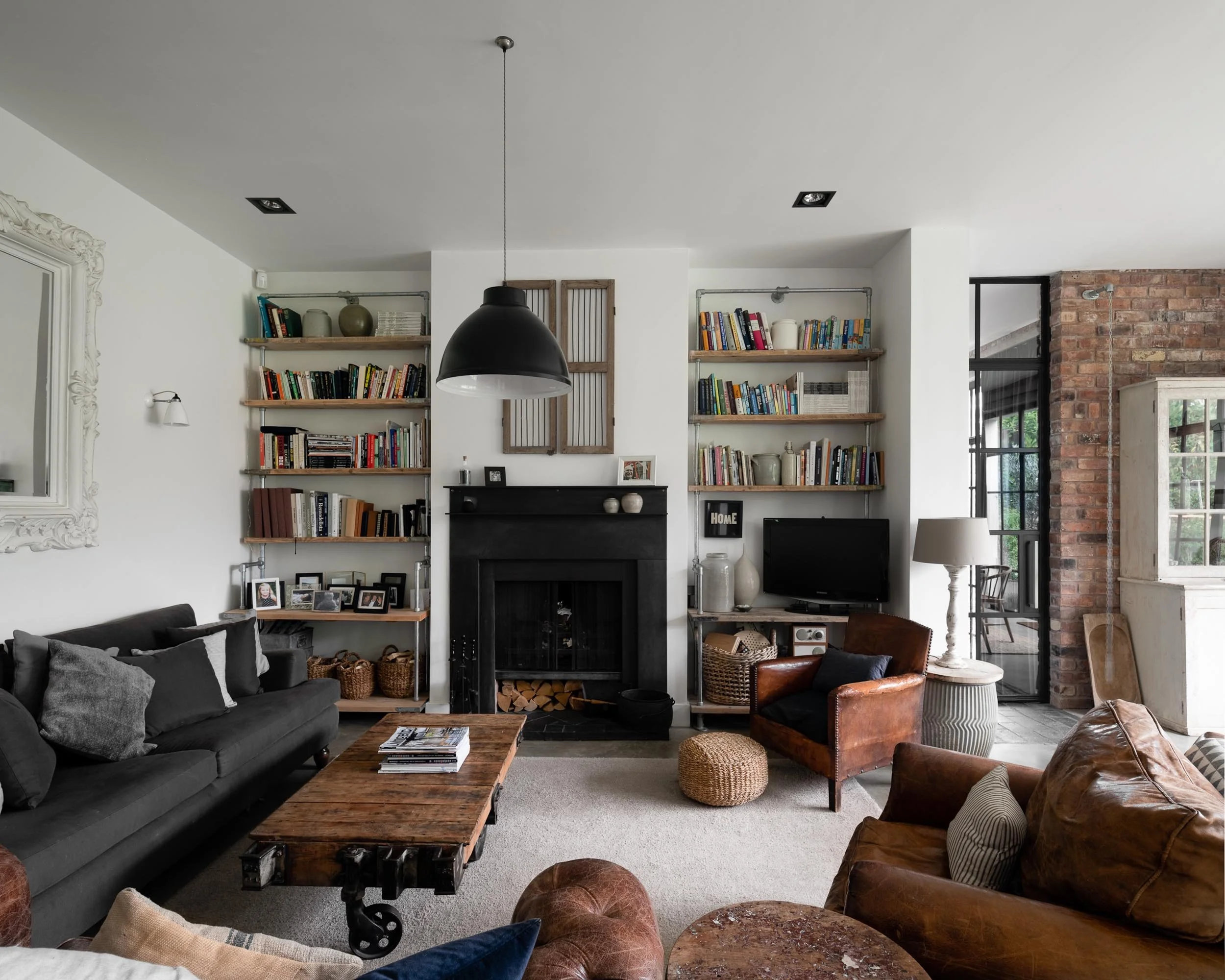 Cozy living room with black sofa, leather armchair, fireplace, wall-mounted shelves with books, decorative baskets, TV, and window with black frame and brick wall in background.