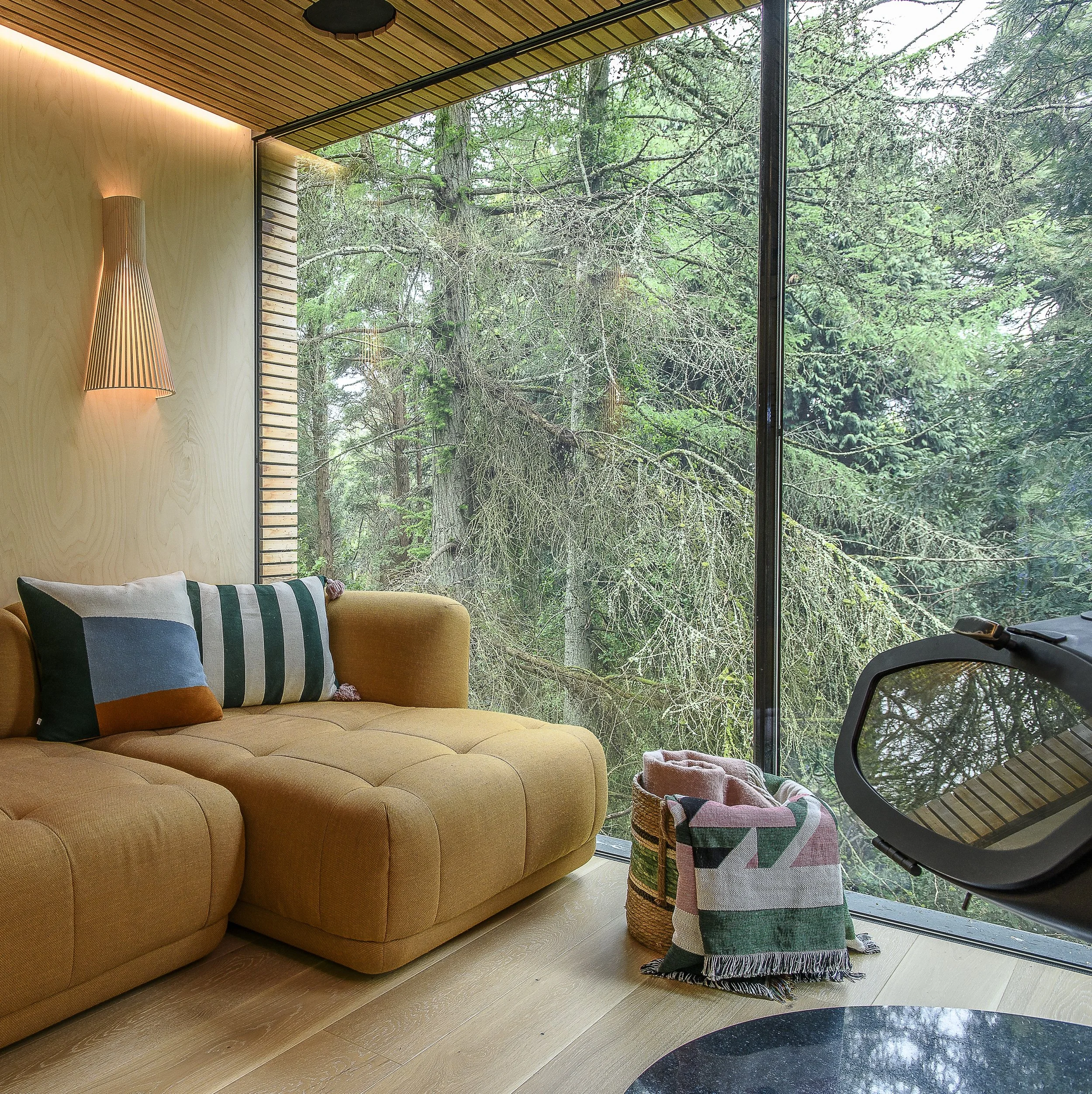 Living room with a mustard-yellow sofa, decorative pillows, a large floor-to-ceiling window showing a view of tall green trees, a wall-mounted lamp, a woven basket with folded blankets, and a virtual reality headset.