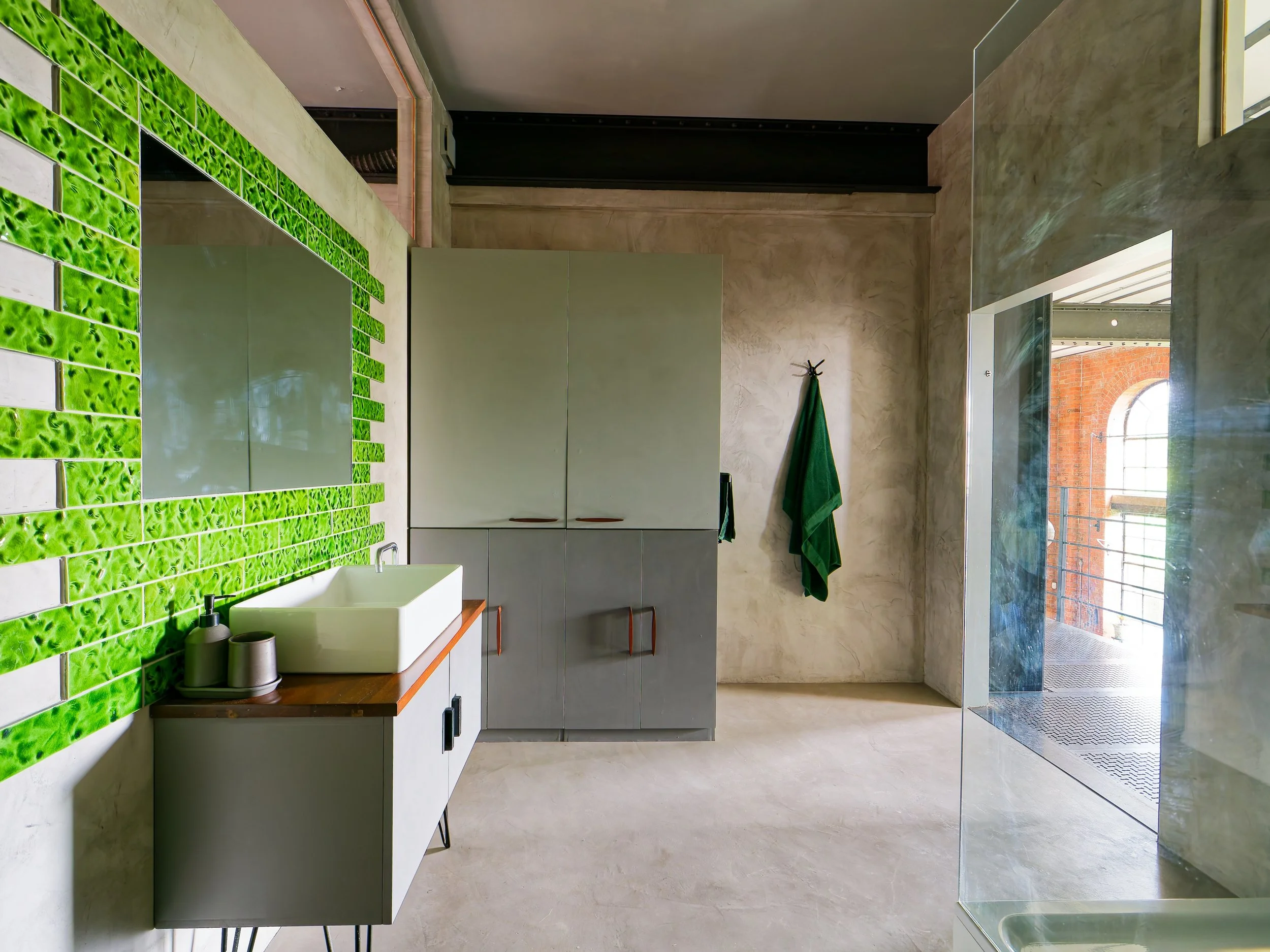 Modern bathroom with green tile wall, white sink on wooden countertop, grey cabinets, a mirror, and a hanging green towel by a windowed door.