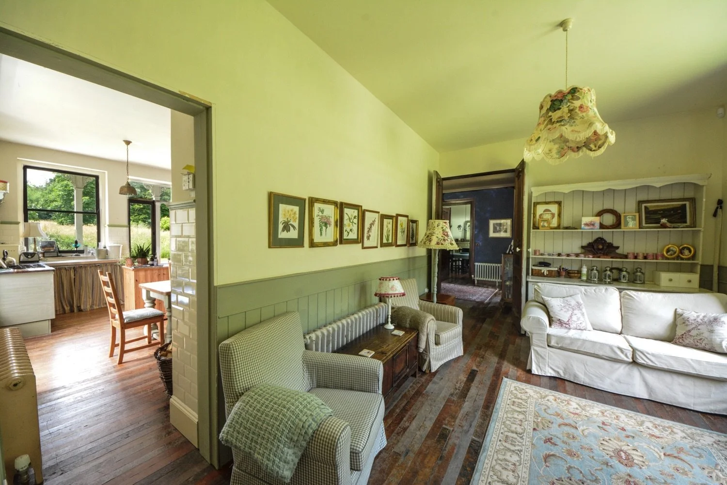 Living room with vintage furniture, floral lampshades, framed artwork, and a white sofa, leading to a kitchen with large windows and outdoor greenery.