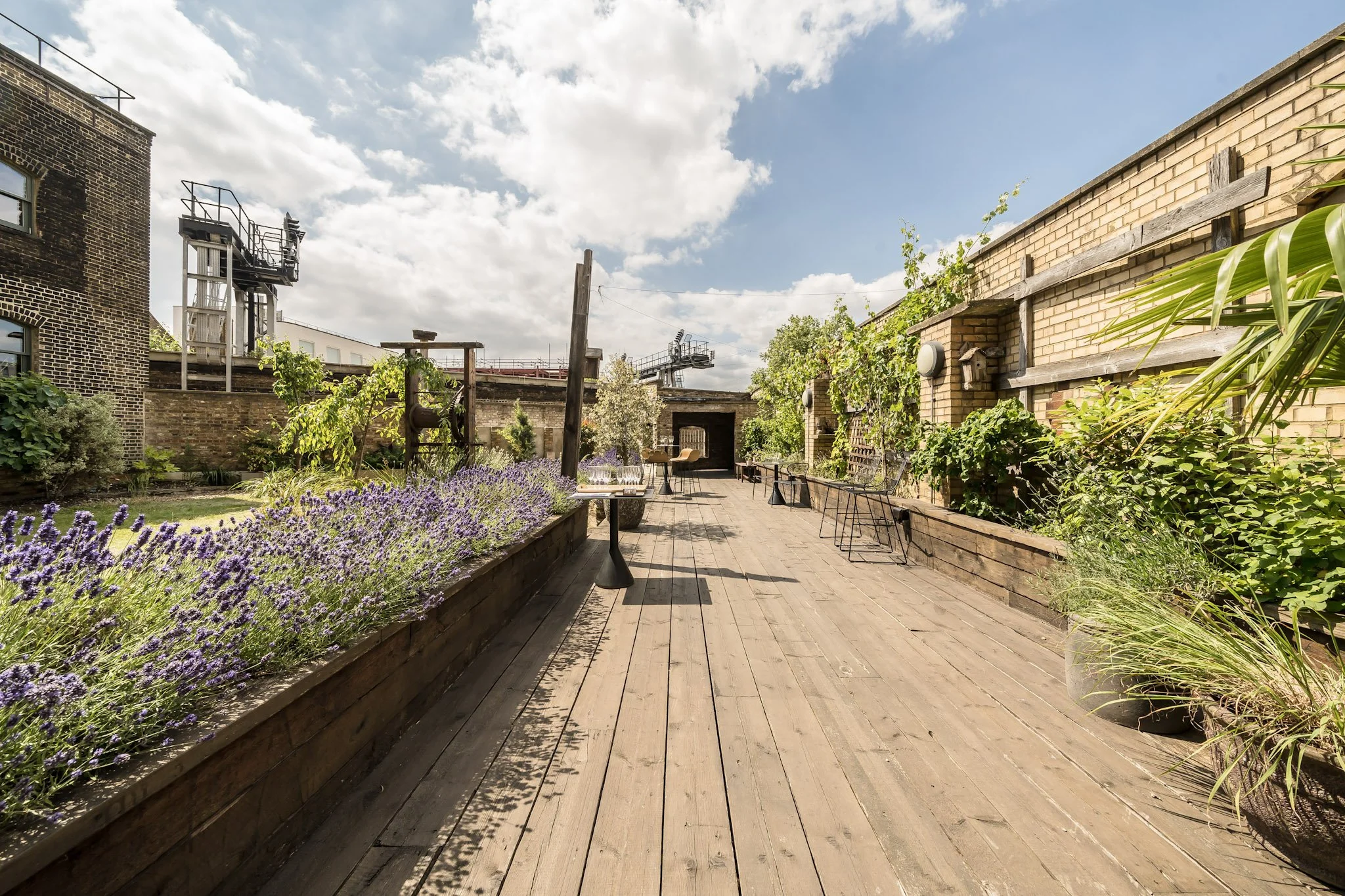 Outdoor rooftop garden with wooden deck, purple lavender plants, potted greenery, seating area with tables and chairs, and brick walls under partly cloudy sky.