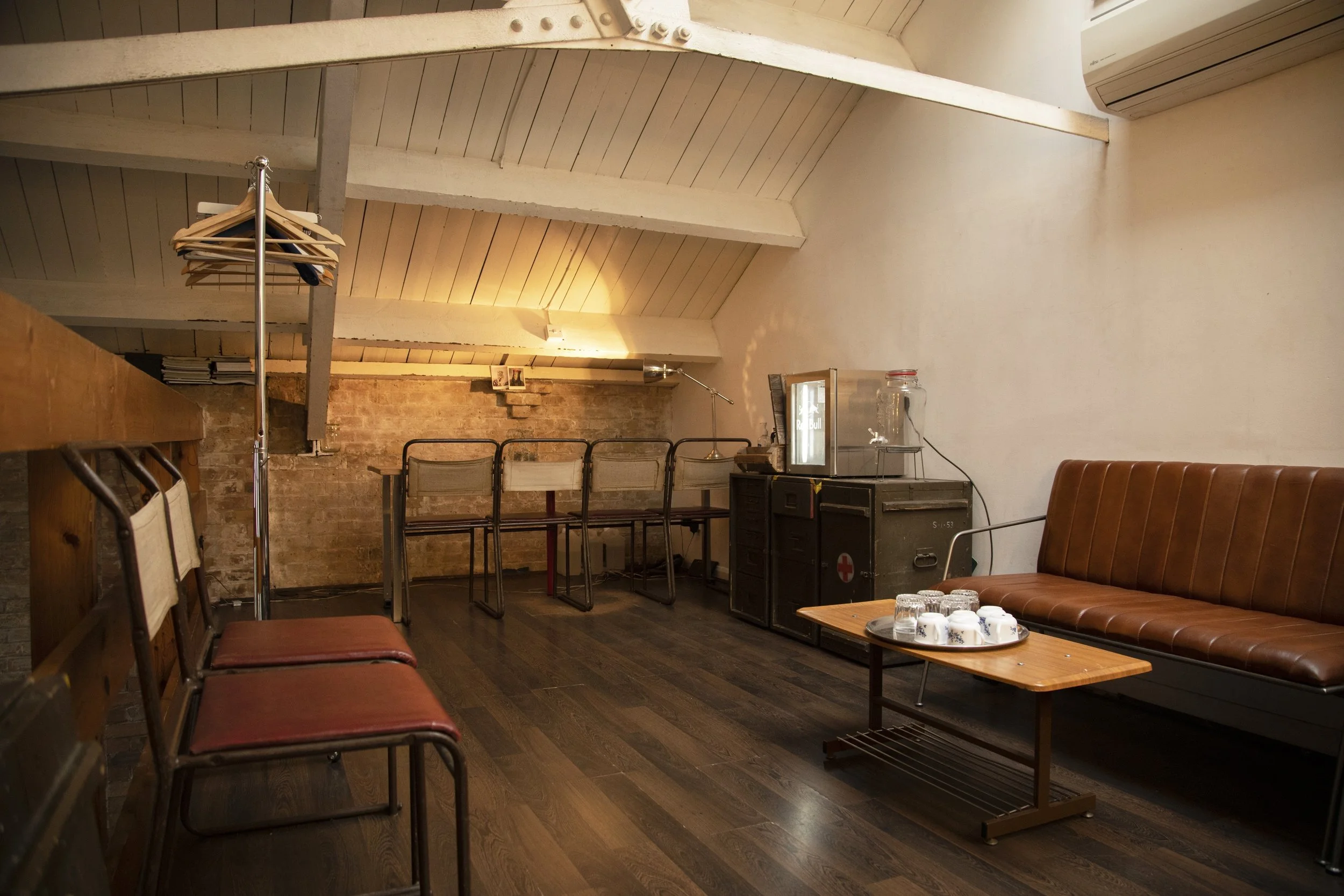 A cozy, vintage-style room with wooden flooring, a brown leather sofa, a wooden coffee table with cups, metal chairs, a small kitchenette, and hanging clothes, with a white-painted sloped ceiling and brick accent wall.