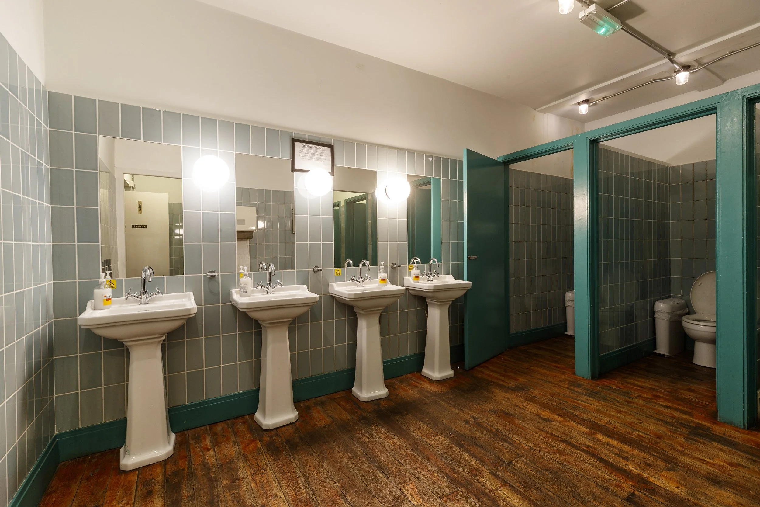 Public restroom with four sinks, mirrors, and soap dispensers on a tiled wall, with stalls containing toilets in the background.