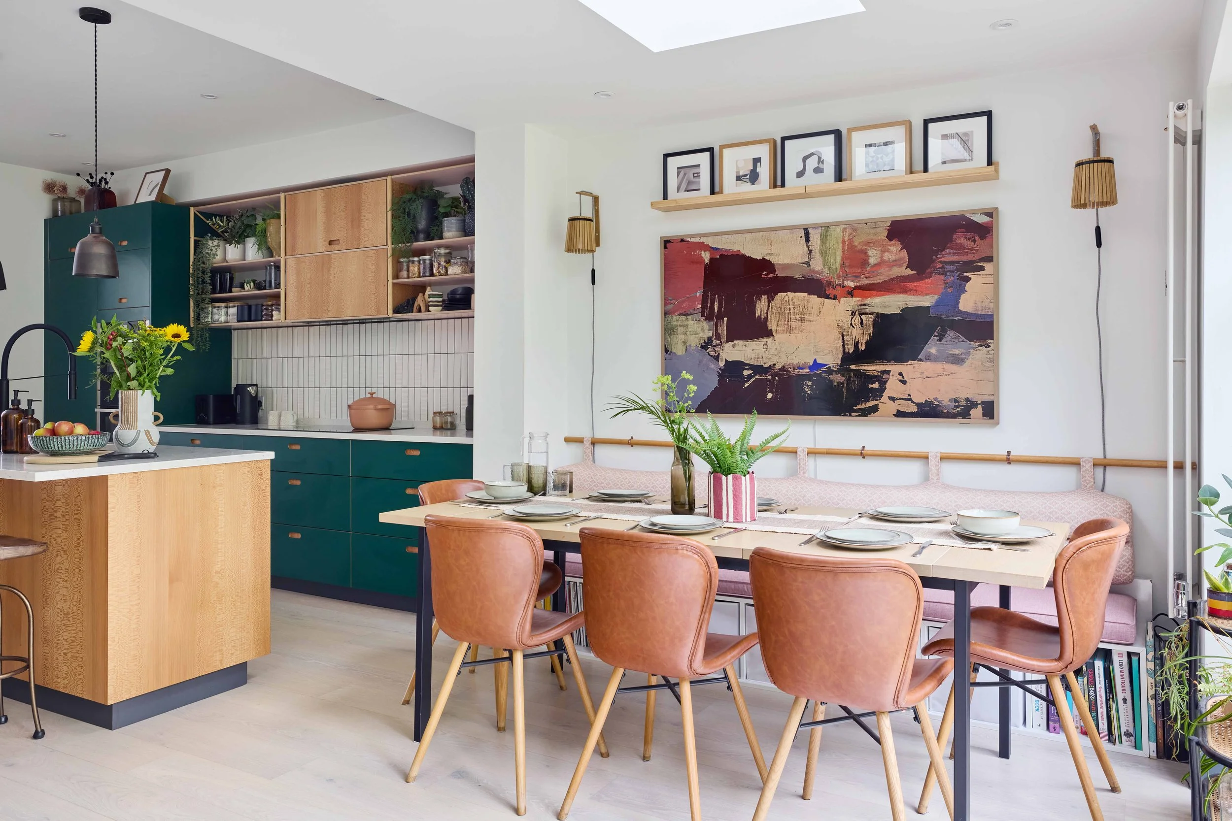Bright modern dining area with a wooden table, pink chairs, and plants, adjacent to a kitchen with green cabinets and open shelving.