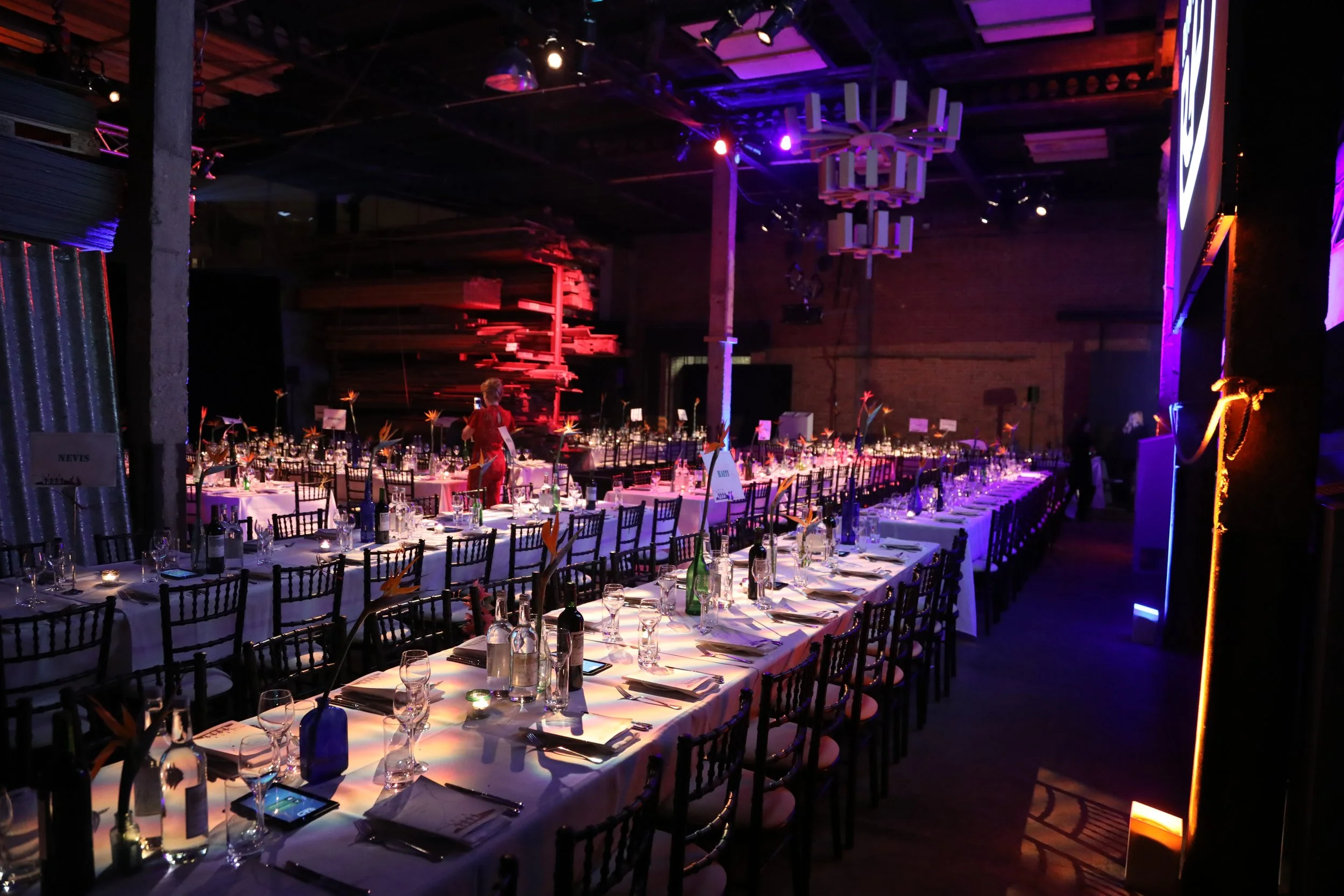 Long banquet tables set with white tablecloths, glassware, and bottle drinks in a dimly lit event hall with purple and red lighting. A person in red attire is on the left side, preparing the tables. The hall features modern decor with a large chandel
