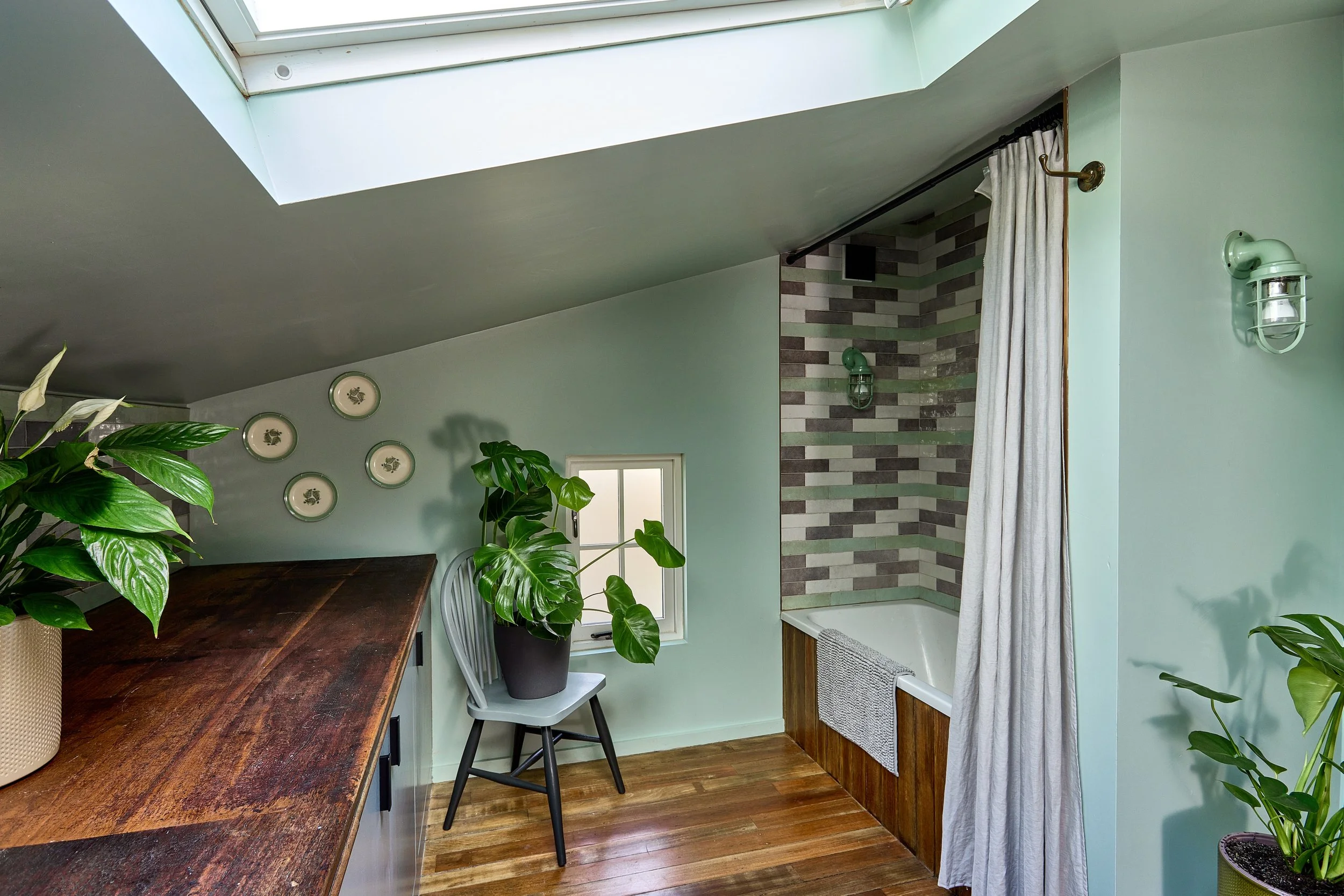 A cozy attic bathroom with a skylight, a small window, wooden floor, a bathtub with a striped tile surround, a curtain, and decorative plates on the wall.