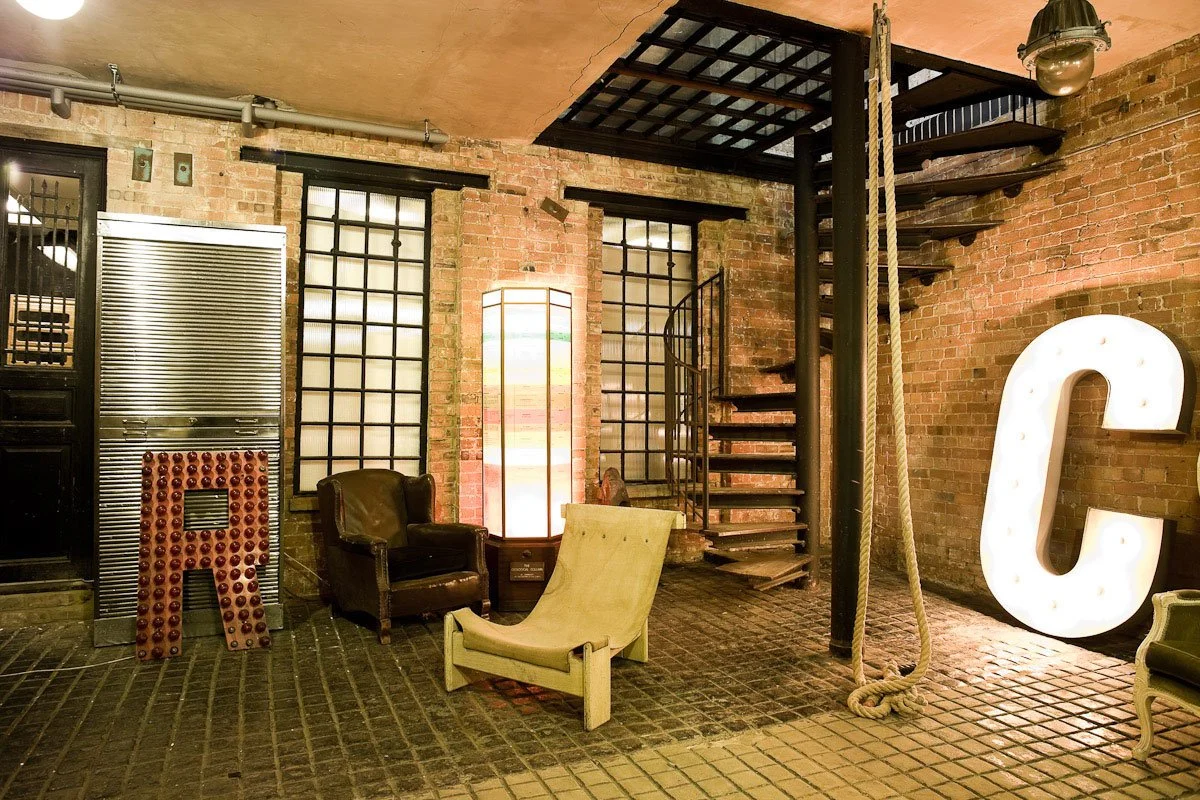 Interior of a brick-walled industrial-style room featuring vintage furniture, a spiral staircase, large illuminated letter C, and other decorative elements.
