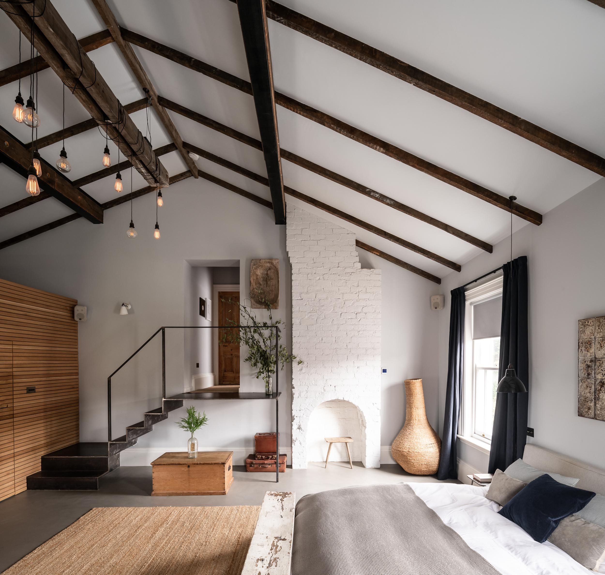 A modern bedroom with a high, vaulted ceiling with exposed dark wood beams and hanging Edison-style light bulbs. The room features a white brick fireplace, black curtains, a large wicker vase, a window, and a bed with gray and white bedding. Decor in