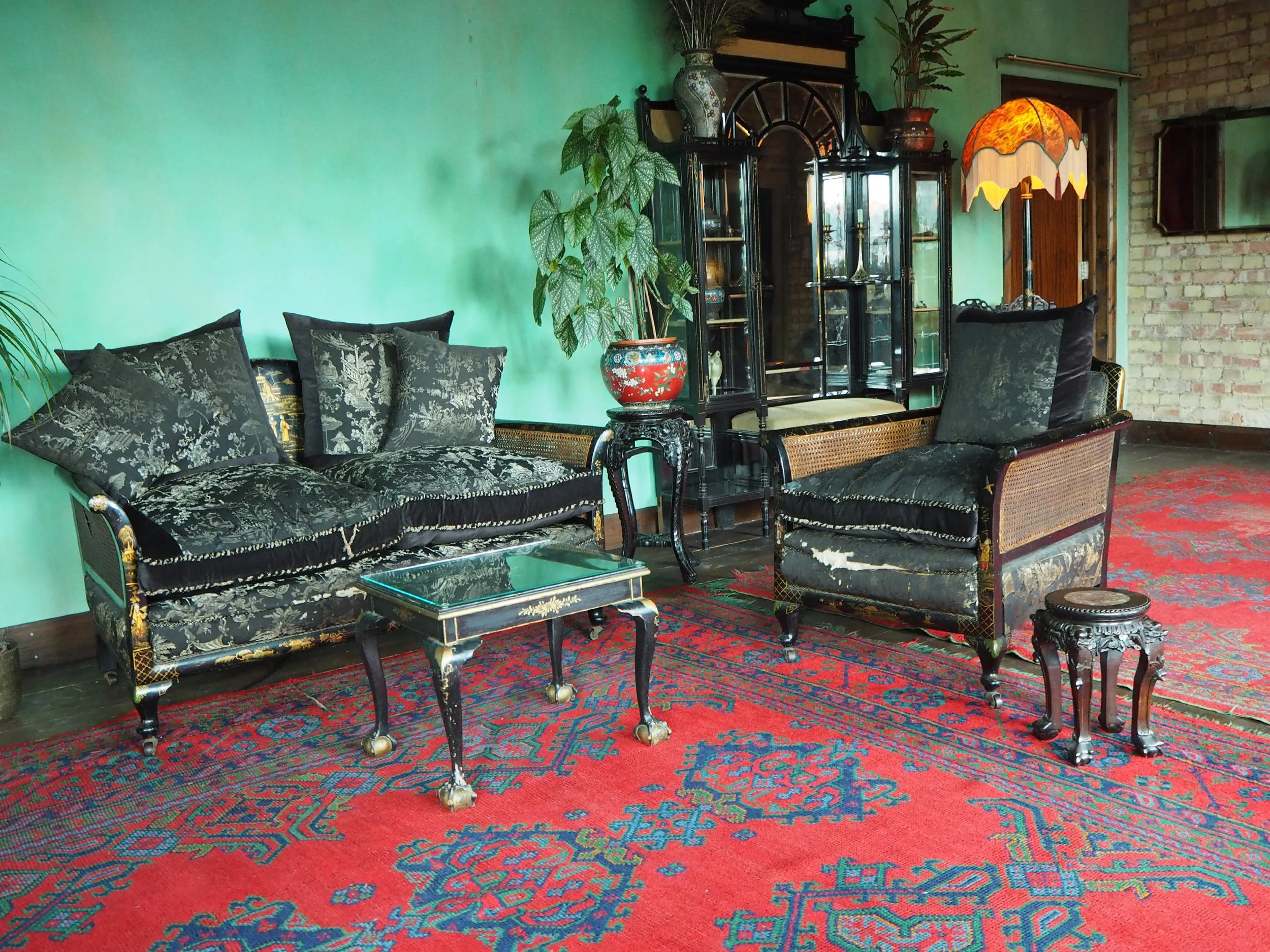 Vintage living room with ornate black and gold upholstered furniture, a large red Persian rug, and a dark glass display cabinet against a green wall.