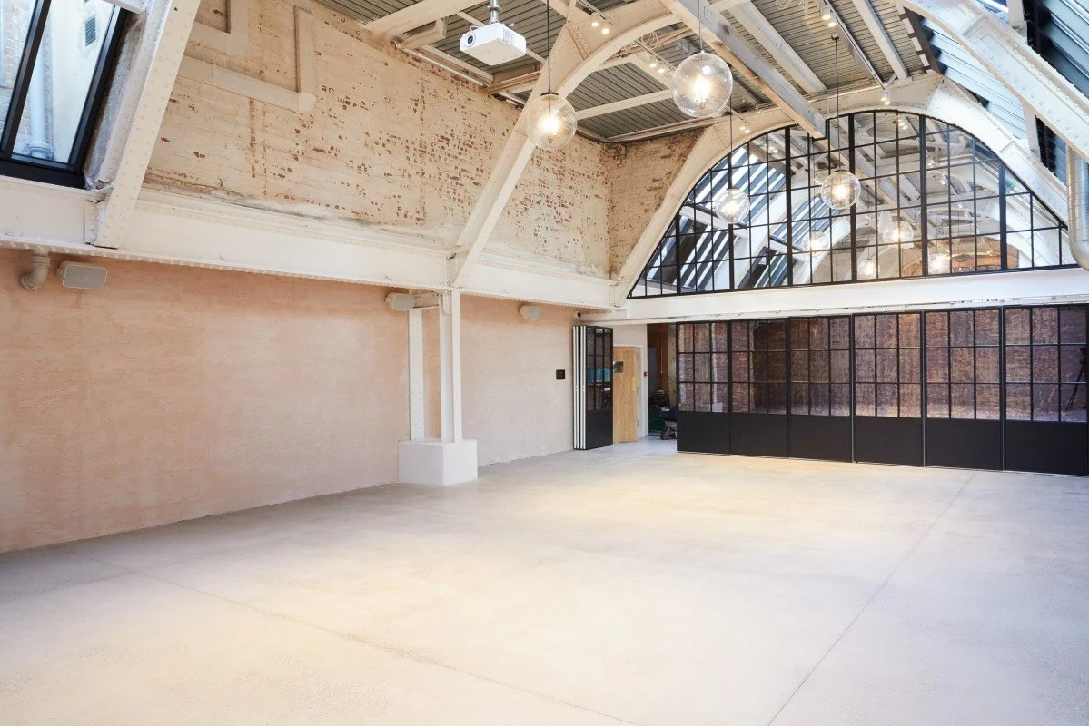 Empty industrial-style room with high ceilings, large arched windows, and pendant lights.