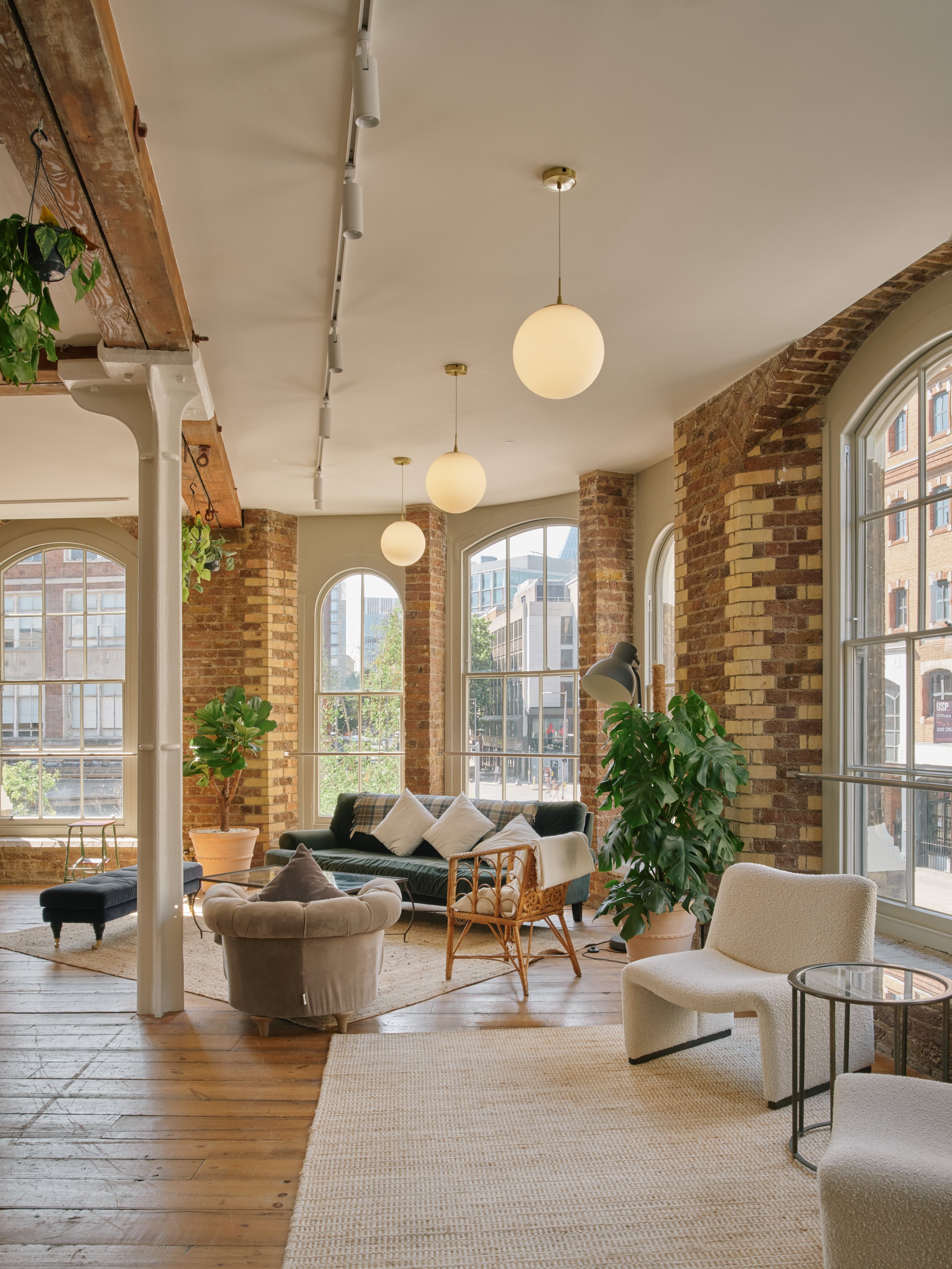 Brightly lit living room with large arched windows, exposed brick walls, and hardwood floors. Features include a dark sofa with white pillows, a white armchair, a wicker chair, a black floor lamp, and indoor plants.