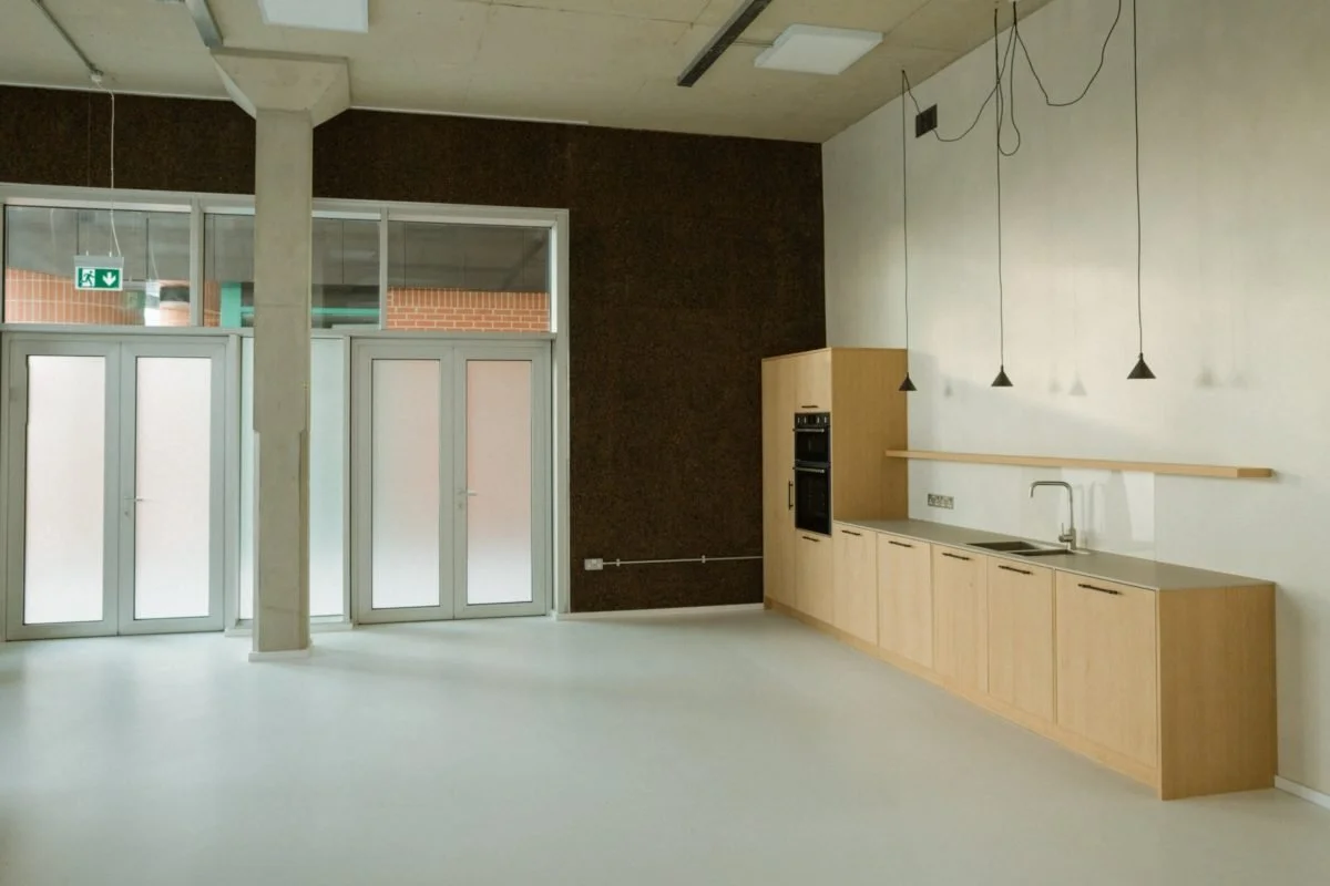 Empty modern kitchen with light wooden cabinets, a sink, and black pendant lights hanging from the ceiling. Large glass doors and a window allow natural light into the space.