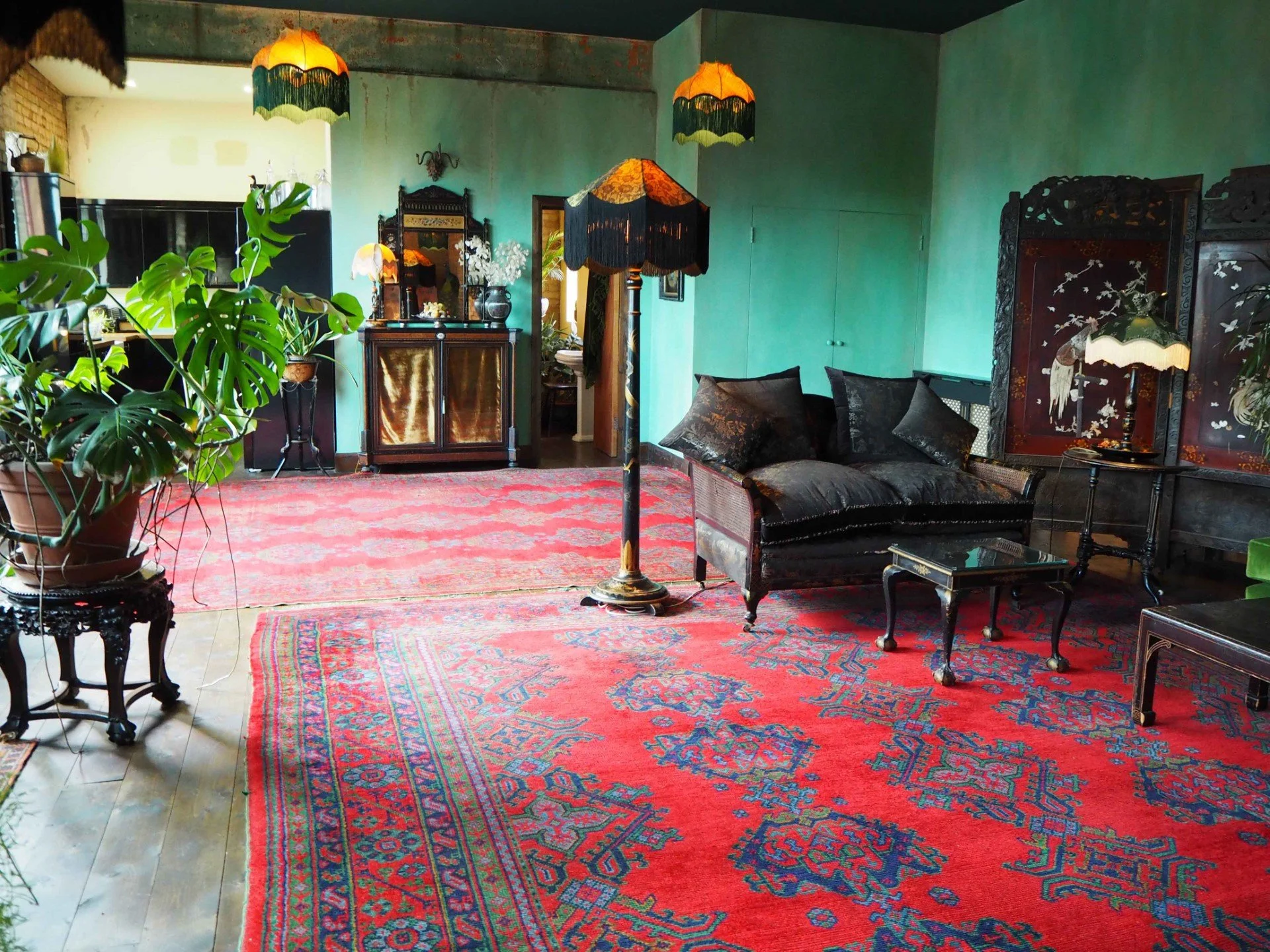 Living room with a large red patterned rug, a black velvet loveseat with black pillows, vintage black side tables, antique lamps with fringed shades, a wooden cabinet, potted plants, and teal walls decorated with artwork.