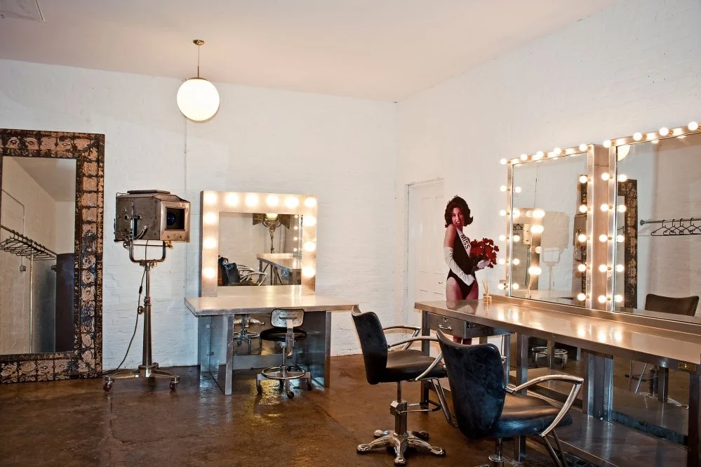 Empty dressing room with large makeup mirrors surrounded by lights, black chairs, a wooden floor, and a woman holding a bouquet of red flowers.