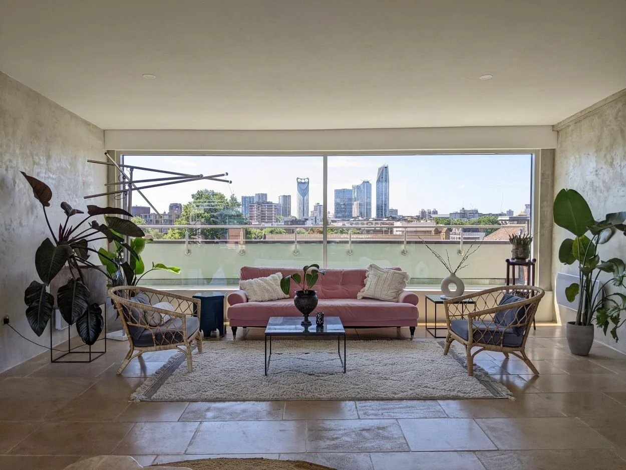 Modern living room with large window overlooking a city skyline. Contains a pink sofa, two wicker chairs with dark cushions, a white rug, and various plants.