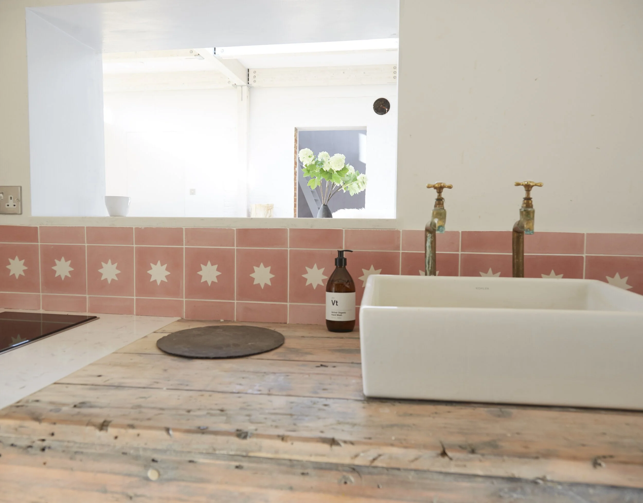 Kitchen sink with pink tiled backsplash, a white farmhouse-style sink, a brown soap dispenser, and a view of a bright open space with green plants in the background.