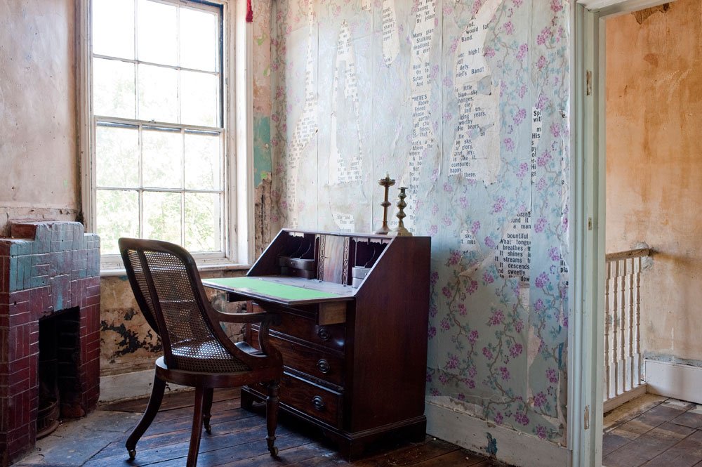 An antique wooden desk with a roll-top, a chair with woven cane back, two candles on top, in a room with peeling painted walls, a large window, and a brick fireplace.