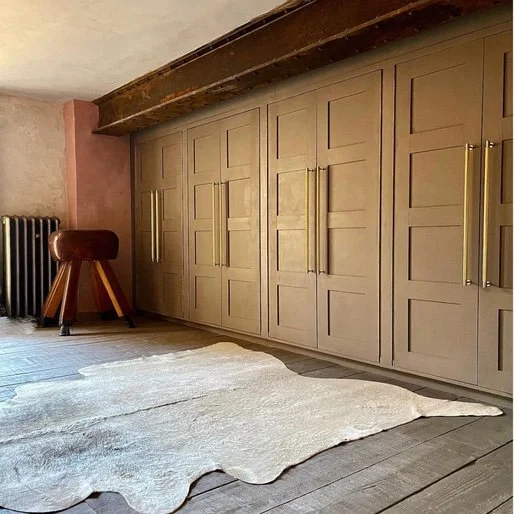 A modern interior room features a large beige built-in wardrobe with gold handles, a wooden beam ceiling, a black radiator, a small round wooden stool, and a light-colored cowhide rug on wooden flooring.