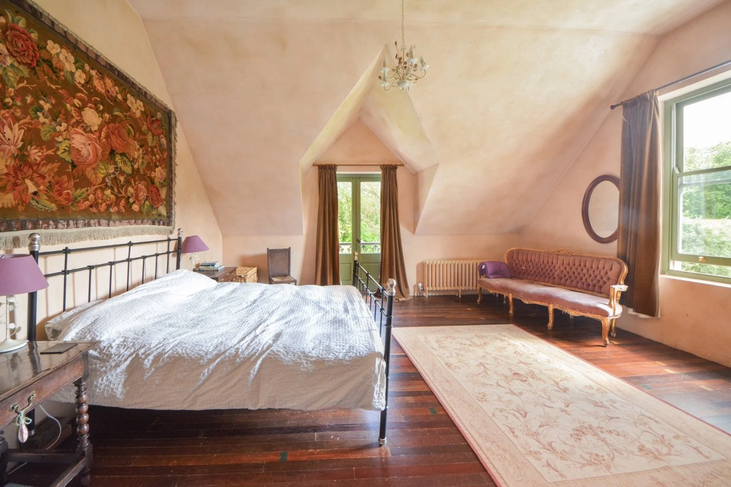 A spacious bedroom with a metal bed frame, white bedding, purple lamps, a tapestry on the wall, a wooden chair, a vintage pink sofa, large windows with curtains, a chandelier, and a wooden floor with a cream-colored rug.