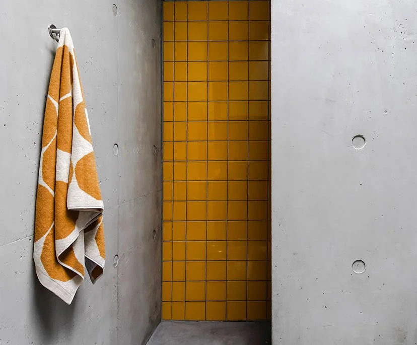 A bathroom corner with a gray concrete wall, an orange and white patterned towel hanging on a metal hook, and a yellow tiled wall.