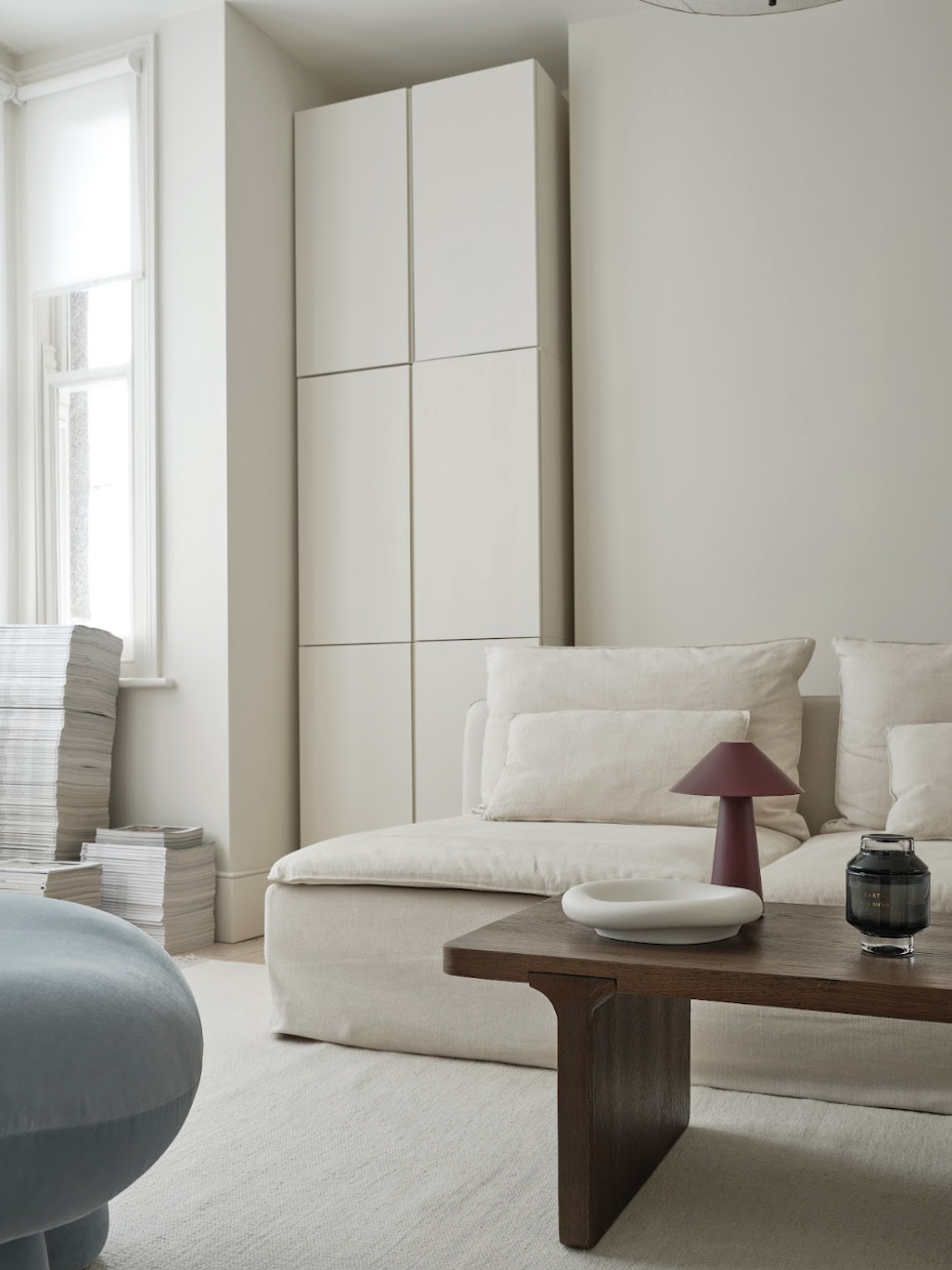 Bright living room with white sofa, wooden coffee table, cream walls, and a tall white cabinet. Stacks of magazines or papers are near the window, which has white blinds.