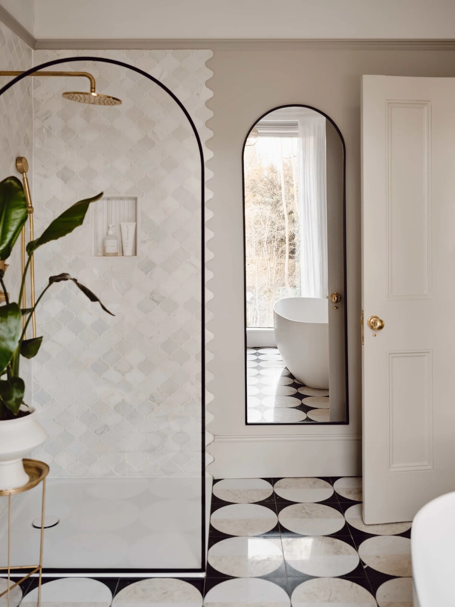 A modern bathroom with a black and white geometric floor, a white freestanding bathtub visible through an open white door, a large oval mirror on the wall, and a partial view of a shower area with a gold showerhead and fixtures, accented by a tiled w