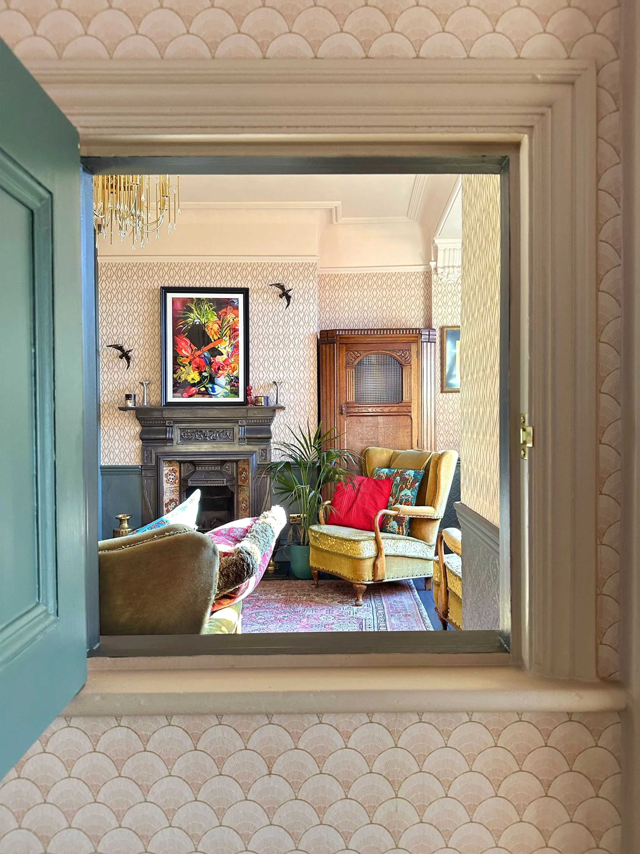 View of a vintage living room through a window with green shutters, featuring a decorative fireplace, colorful artwork, chandelier, three upholstered armchairs with colorful pillows, bookshelf, potted plant, patterned wallpaper, and a rug.