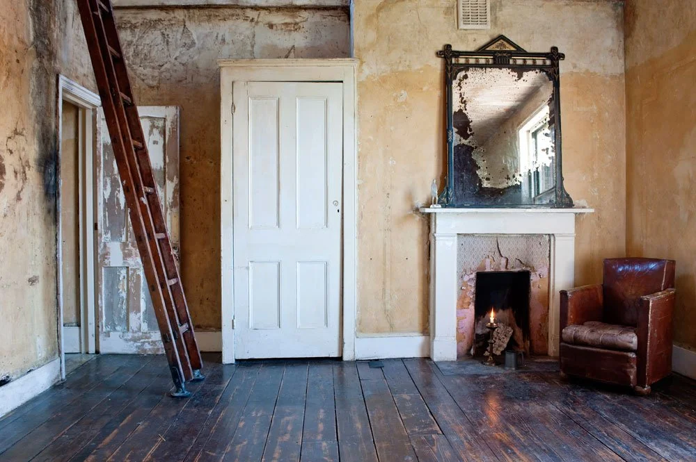 Old room with peeling yellow walls, an open door, a tall ladder leaning against the wall, a white fireplace with a mirror above it, a small fire burning inside, and a worn leather armchair to the right.