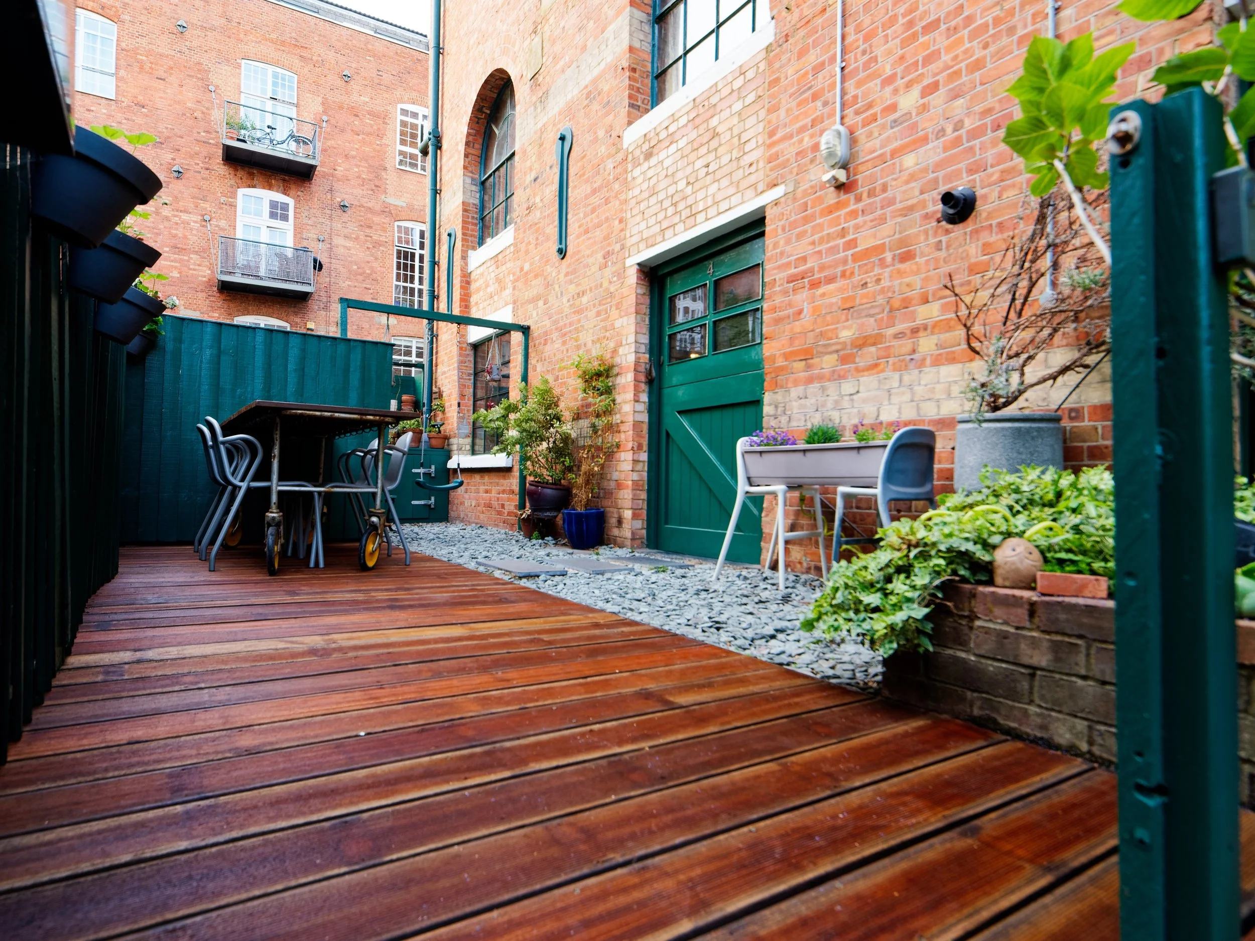 Urban backyard with wooden deck, green door, potted plants, outdoor furniture, and brick walls.