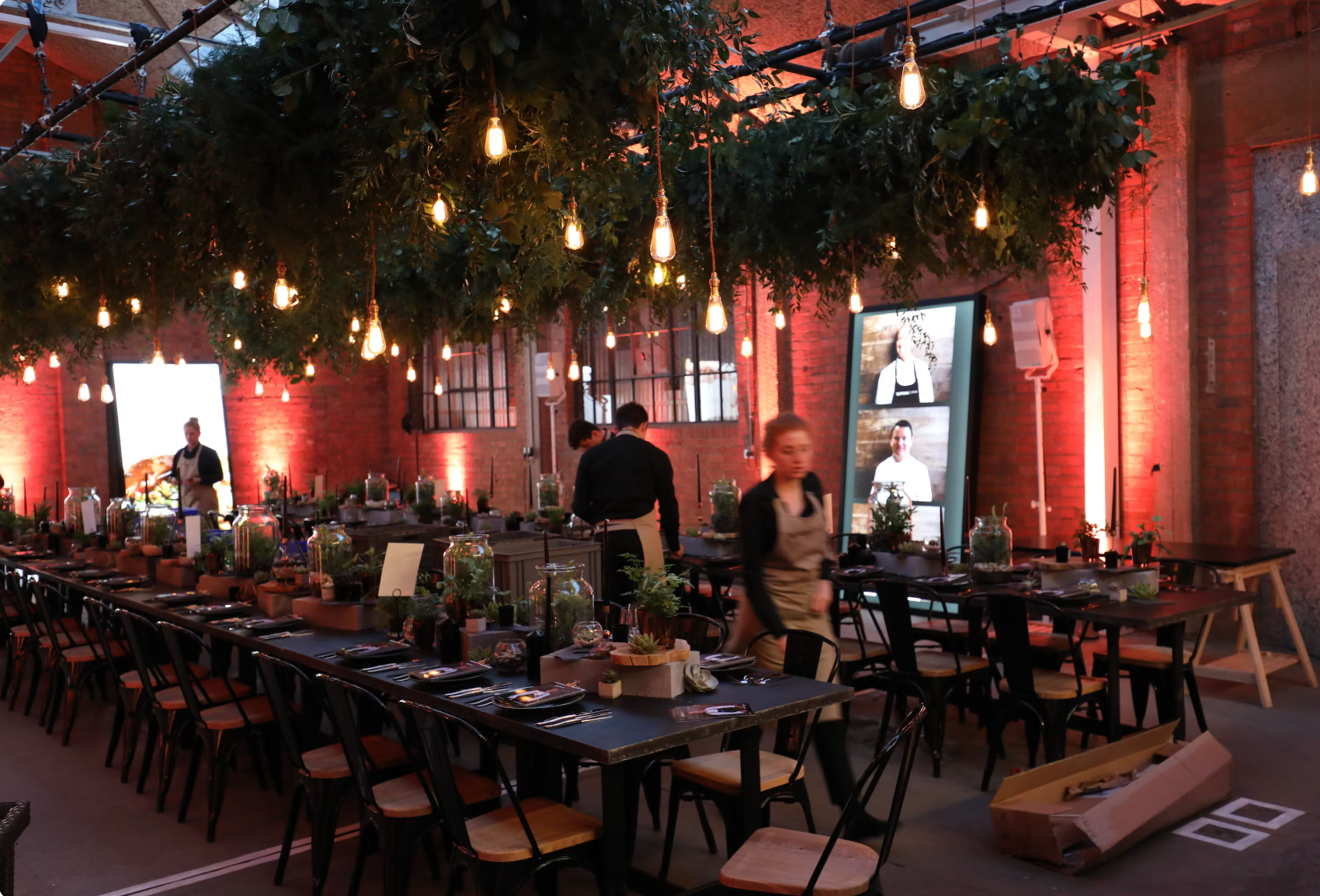 Inside a restaurant with brick walls and hanging lights, staff preparing tables decorated with plants and tableware.