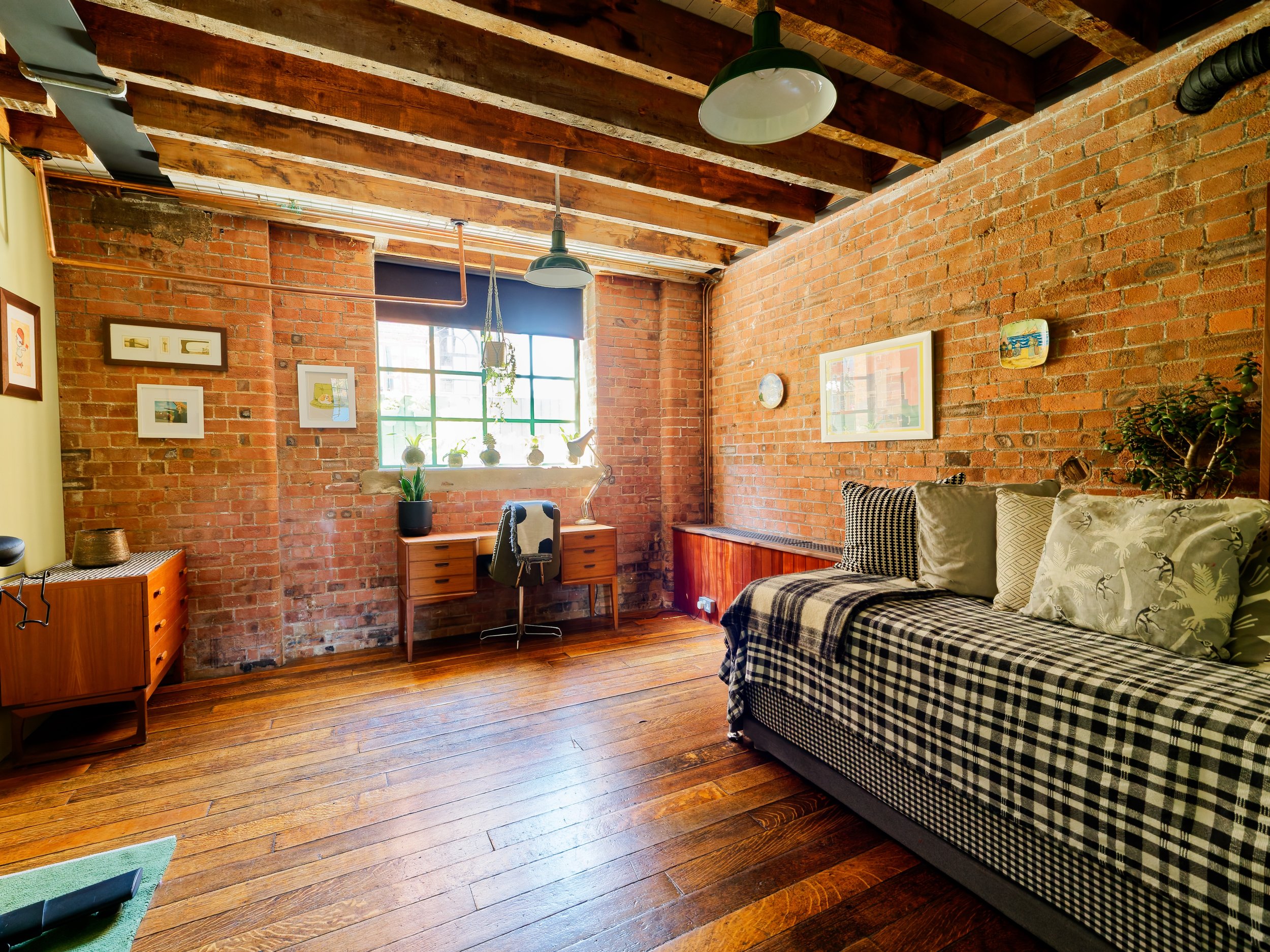 A cozy living room with exposed brick walls, wooden flooring, a window with plants, a black and white checkered sofa with pillows, a wooden desk with a swivel chair, and hanging industrial-style lights.