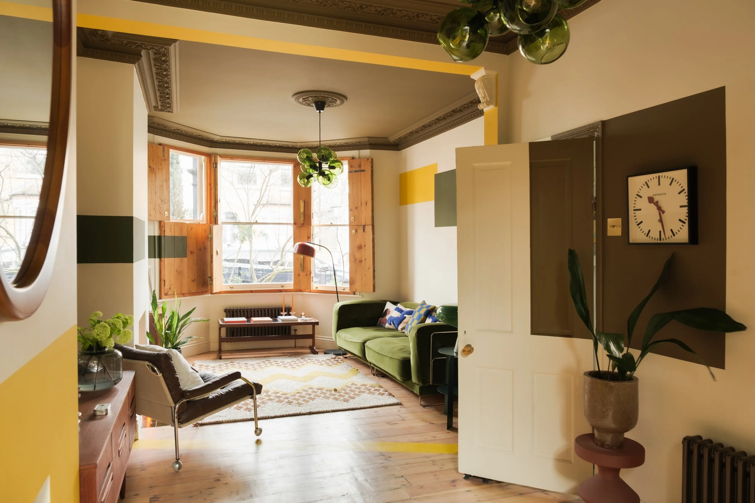 A cozy living room with a large bay window, green sofa, brown armchair, plants, a geometric rug, and a ceiling light fixture with green glass globes.