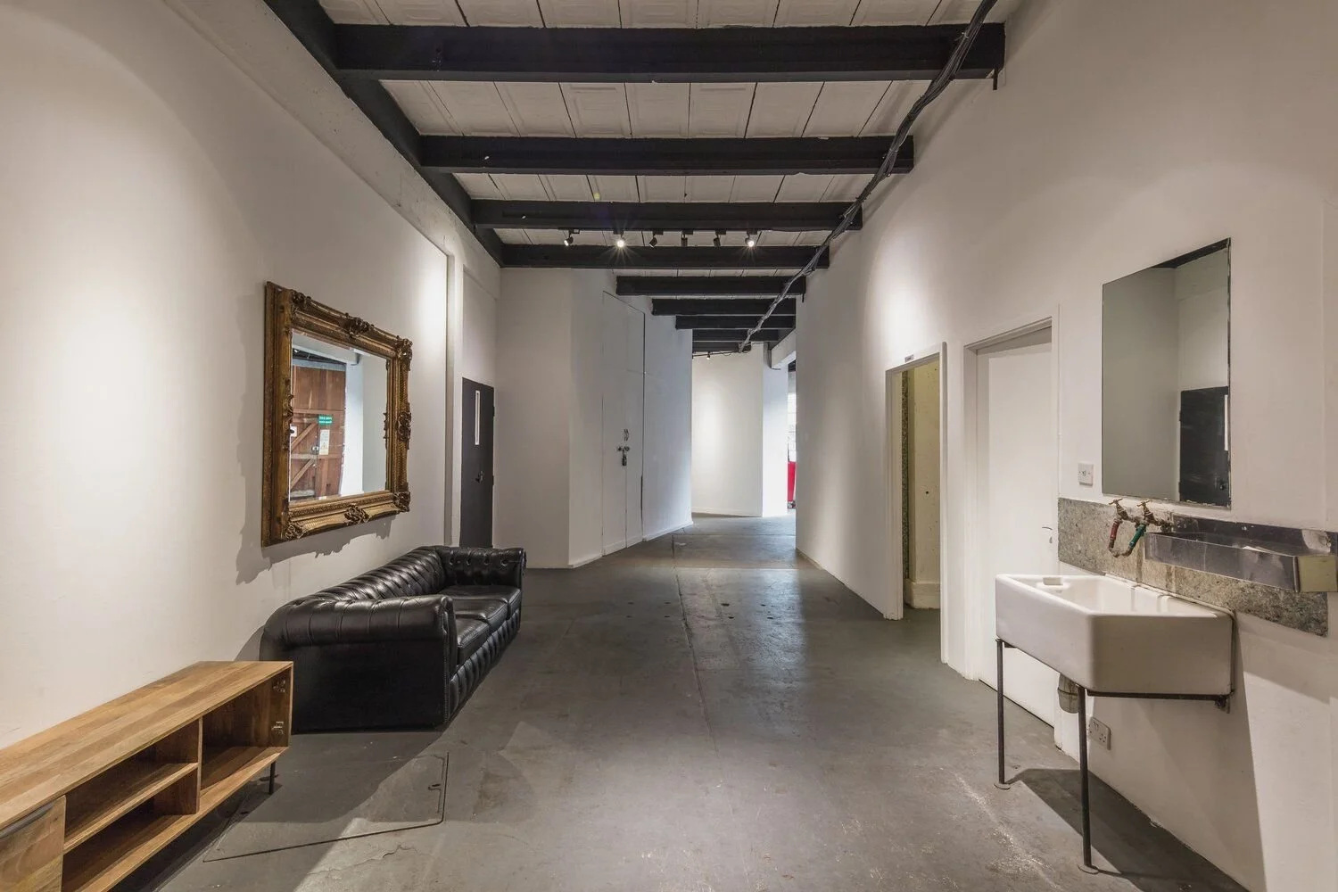 Interior of an industrial-style room with white walls, black ceiling beams, and concrete flooring. There is a black leather couch, a wooden shelf, a framed mirror on the wall, a white utility sink, and doorway openings along the hallway.