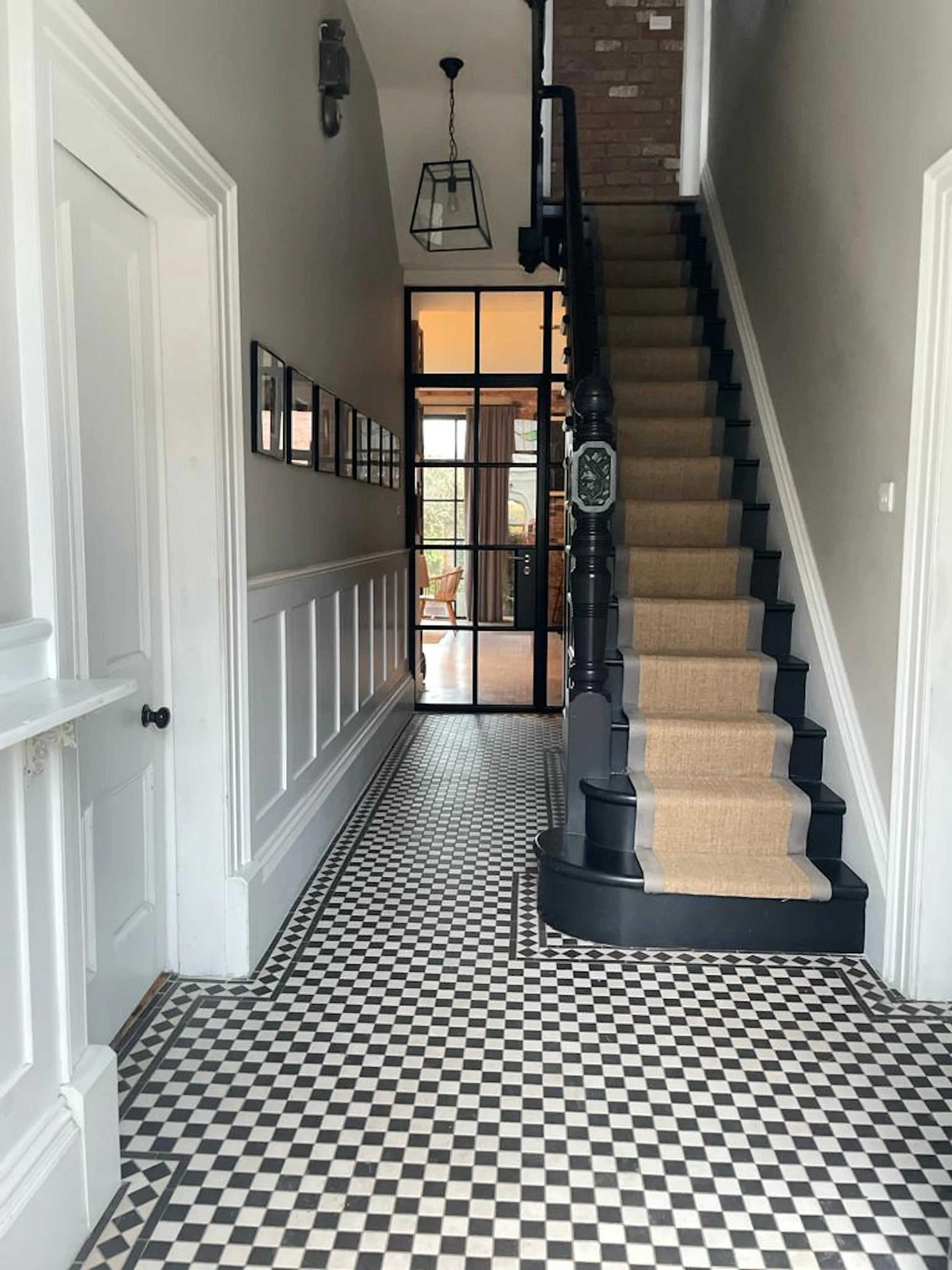 Interior entryway with black and white checkered tile floor, staircase with beige carpet runner, black balustrade, and glass-paneled door leading to another room with wooden furniture and large windows.