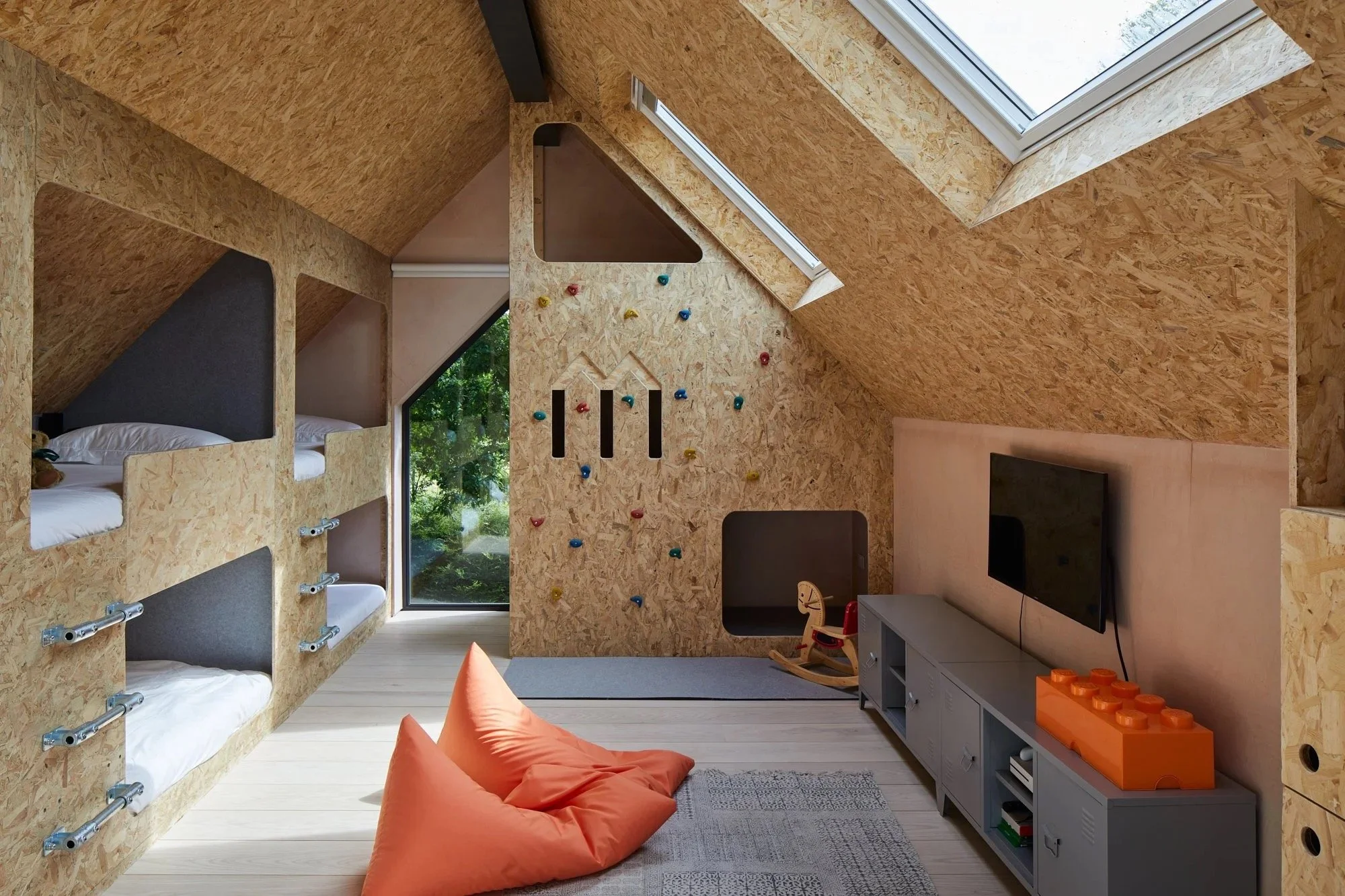 Interior of a modern child's bedroom with a unique sloped ceiling, featuring built-in beds, a climbing wall, a TV, and play area with a rocking horse and bean bag chairs.