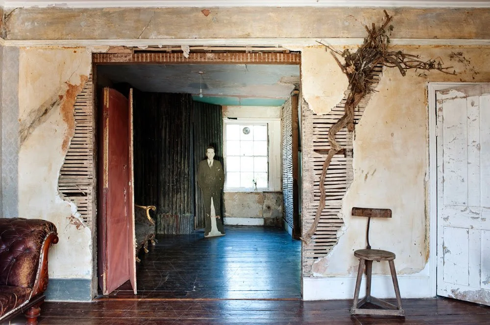 An interior room with a partially torn wall revealing a neighbor room with a cardboard cutout of a man standing near the window. The room has a wooden floor, a vintage chair, a wooden chair, and old, peeling walls.