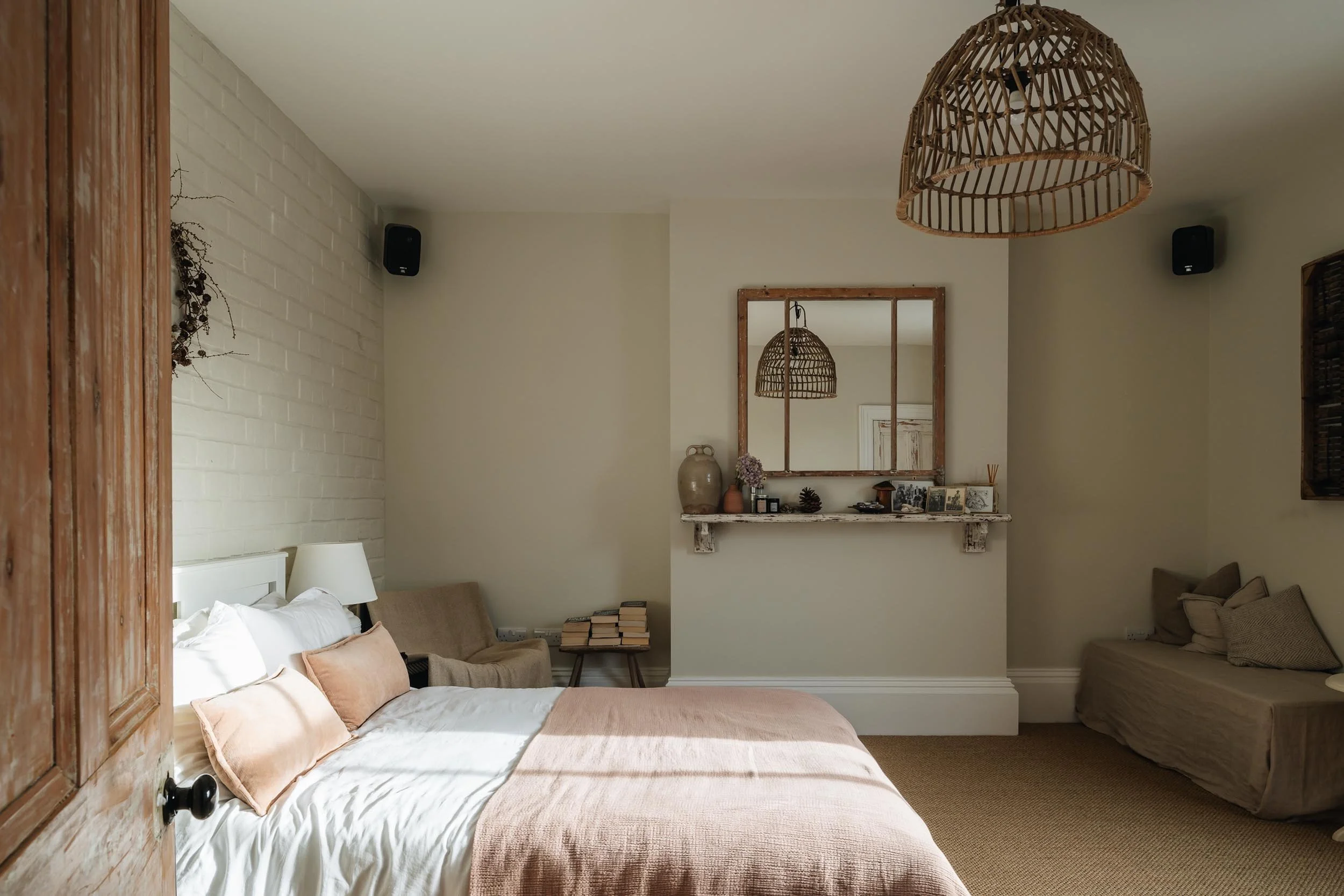 A cozy bedroom with a white bed, beige pillows, a beige bench, a side table with books, and a sitting area with cushions, a white brick wall, a wooden wardrobe, and decorative items on a shelf, with a mirror reflecting a wicker lampshade.