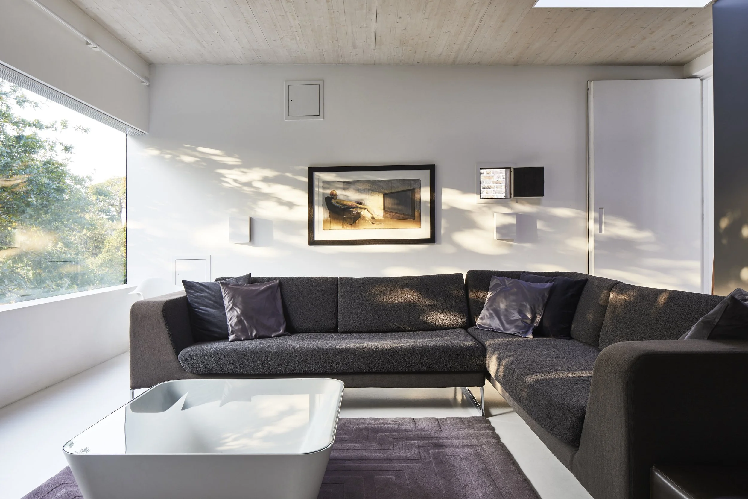 Modern living room with a large L-shaped gray sofa, purple pillows, glass coffee table, with a framed painting and white wall-mounted lights, sunlight streaming through a large window.
