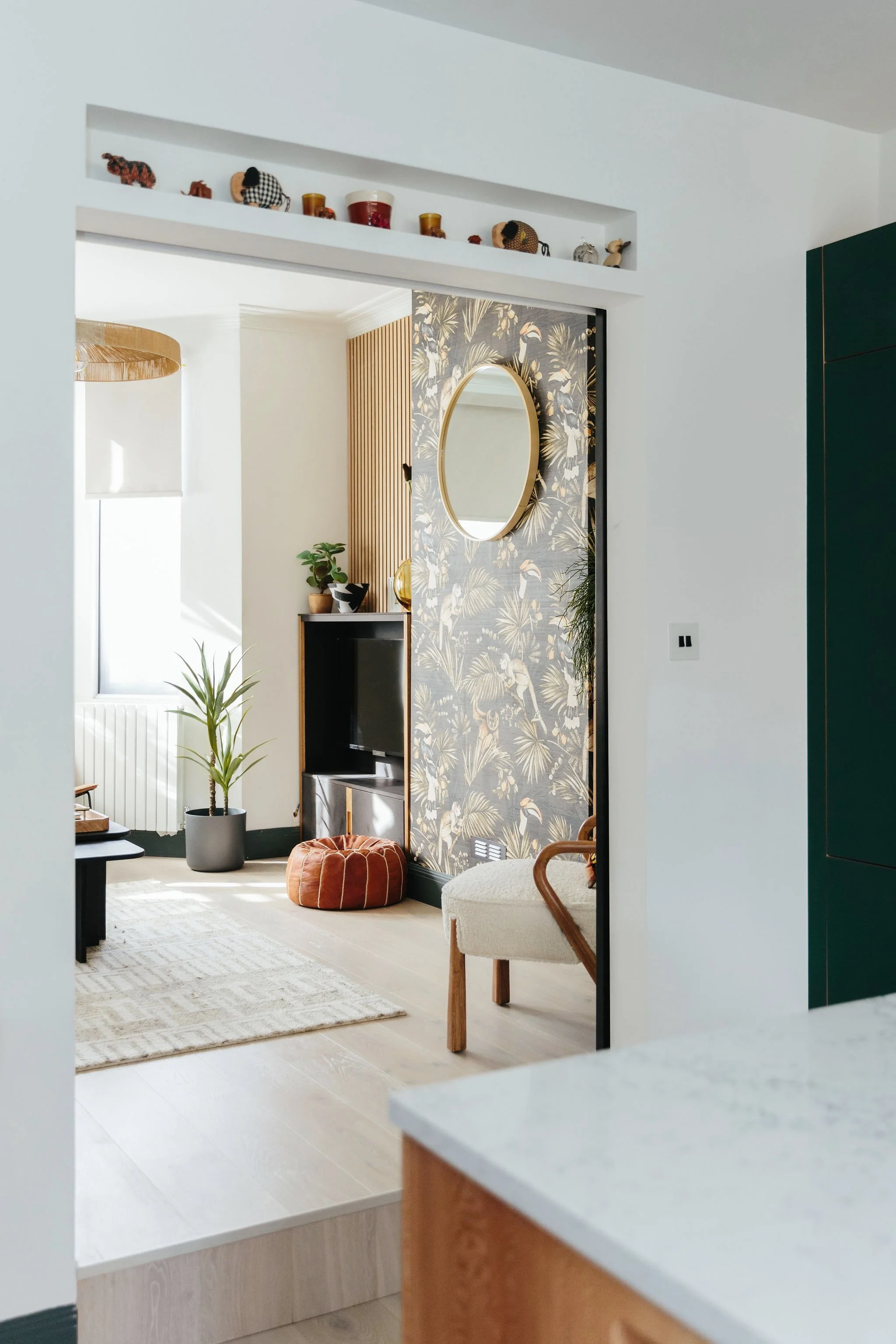 Living room with tropical patterned wallpaper, mirror, plants, TV, ottoman, and modern furniture.