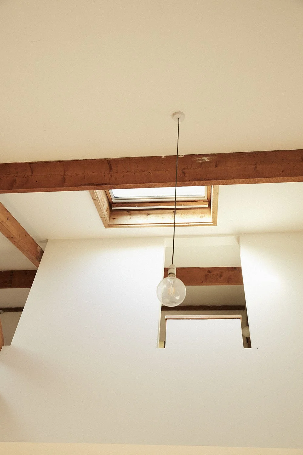 Indoor ceiling with a hanging lightbulb, wooden beams, and a ceiling window letting in natural light.